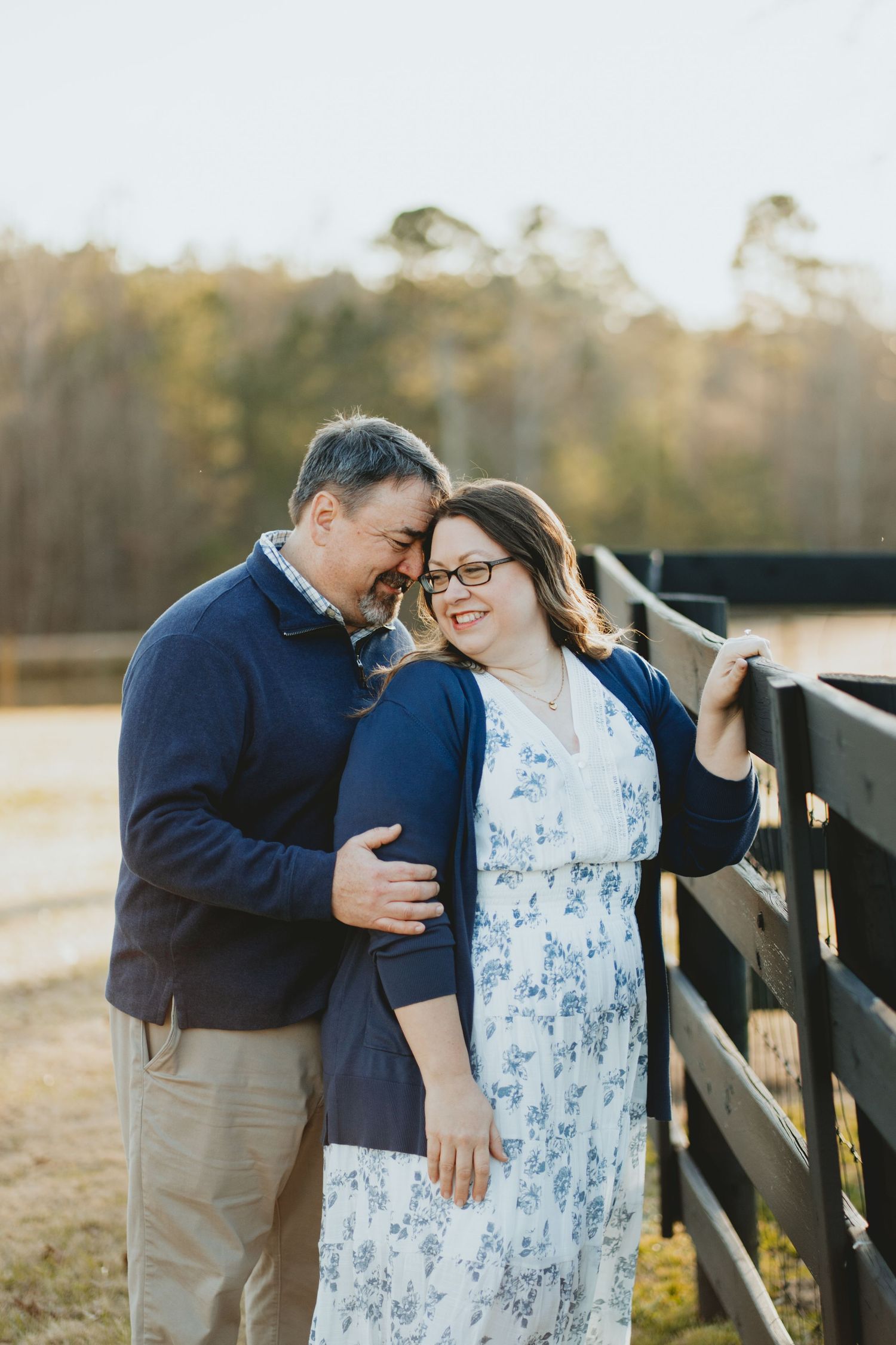 Smiling couple leaning against a wooden fence at golden hour on a farm property.