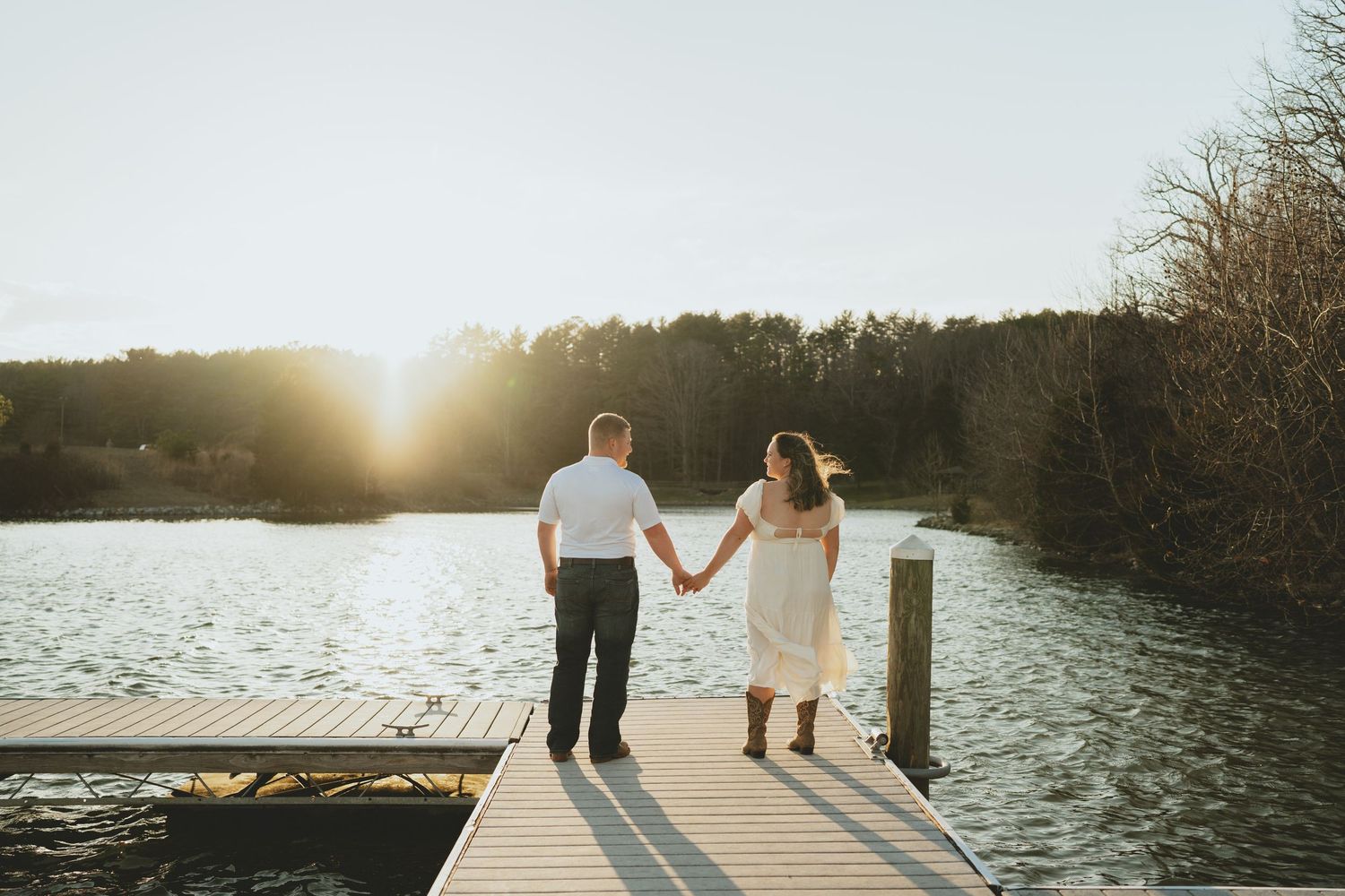 Couple holding hands walking toward a glowing sunset on a wooden dock over a calm lake.