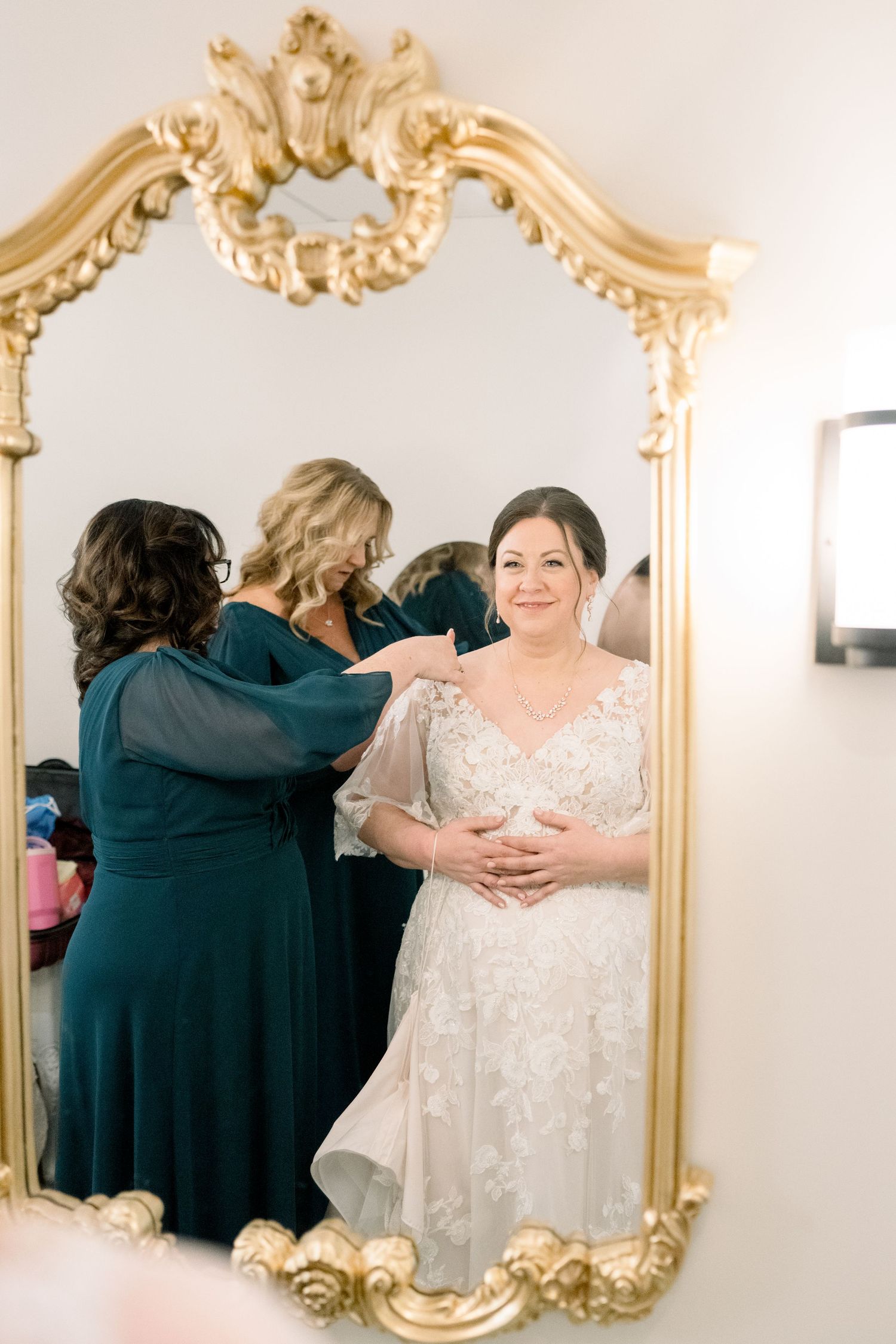 Bride smiles in gold ornate mirror as bridesmaids in teal dresses help her into a lace wedding gown.
