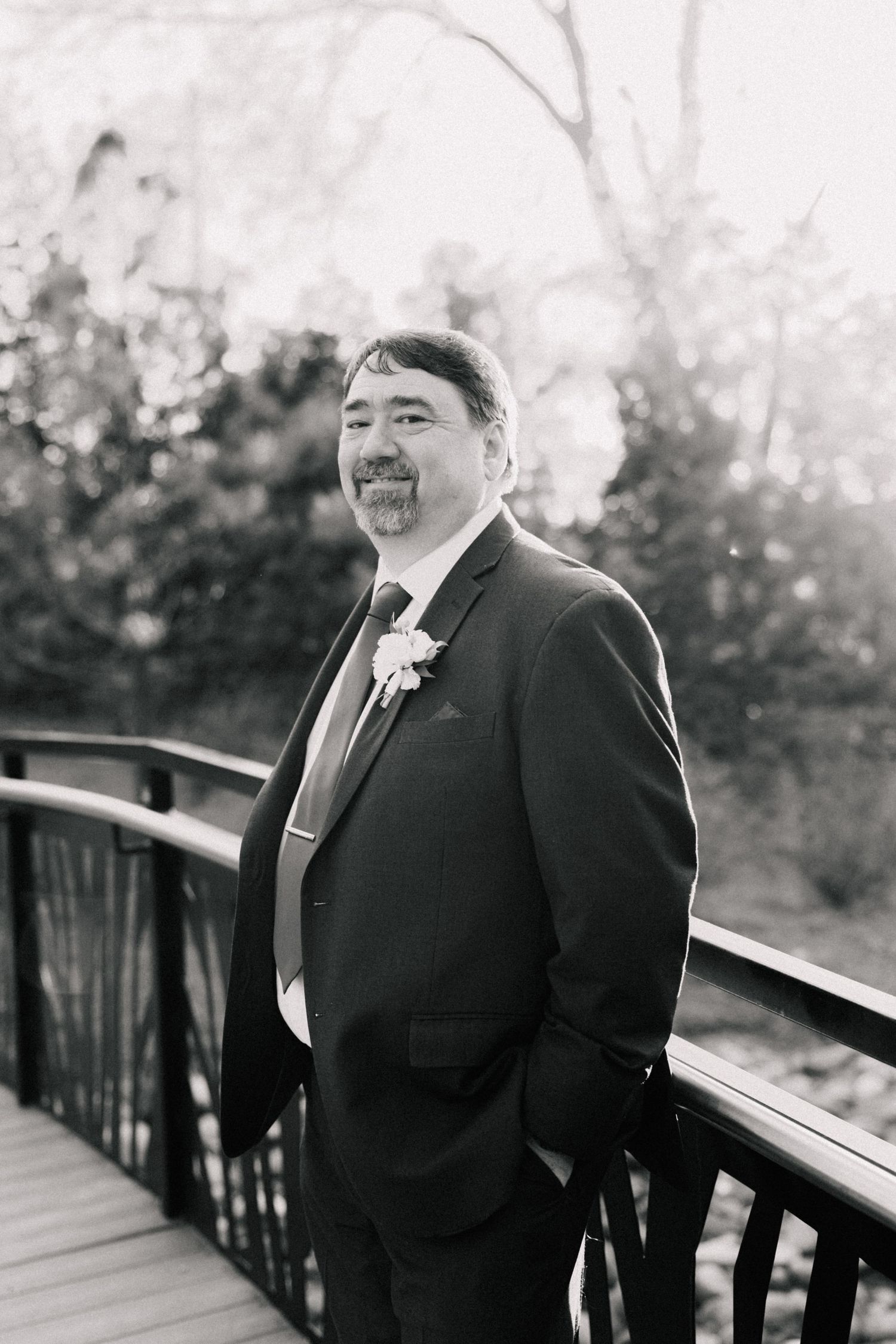 Handsome groom in dark suit with boutonniere posing on wooden bridge in black and white.