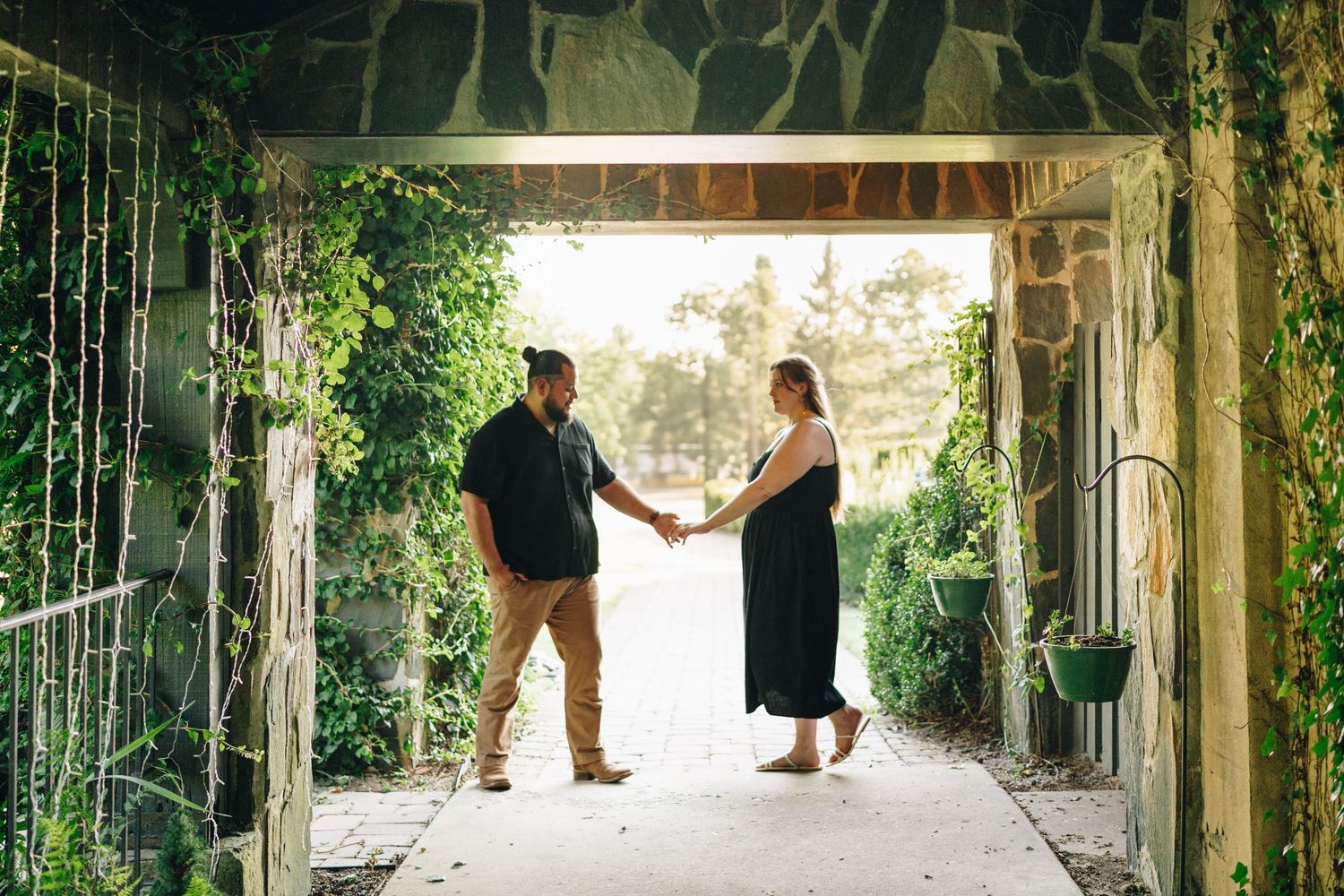 Couple holding hands facing each other in an ivy-covered stone archway passage with green plants.