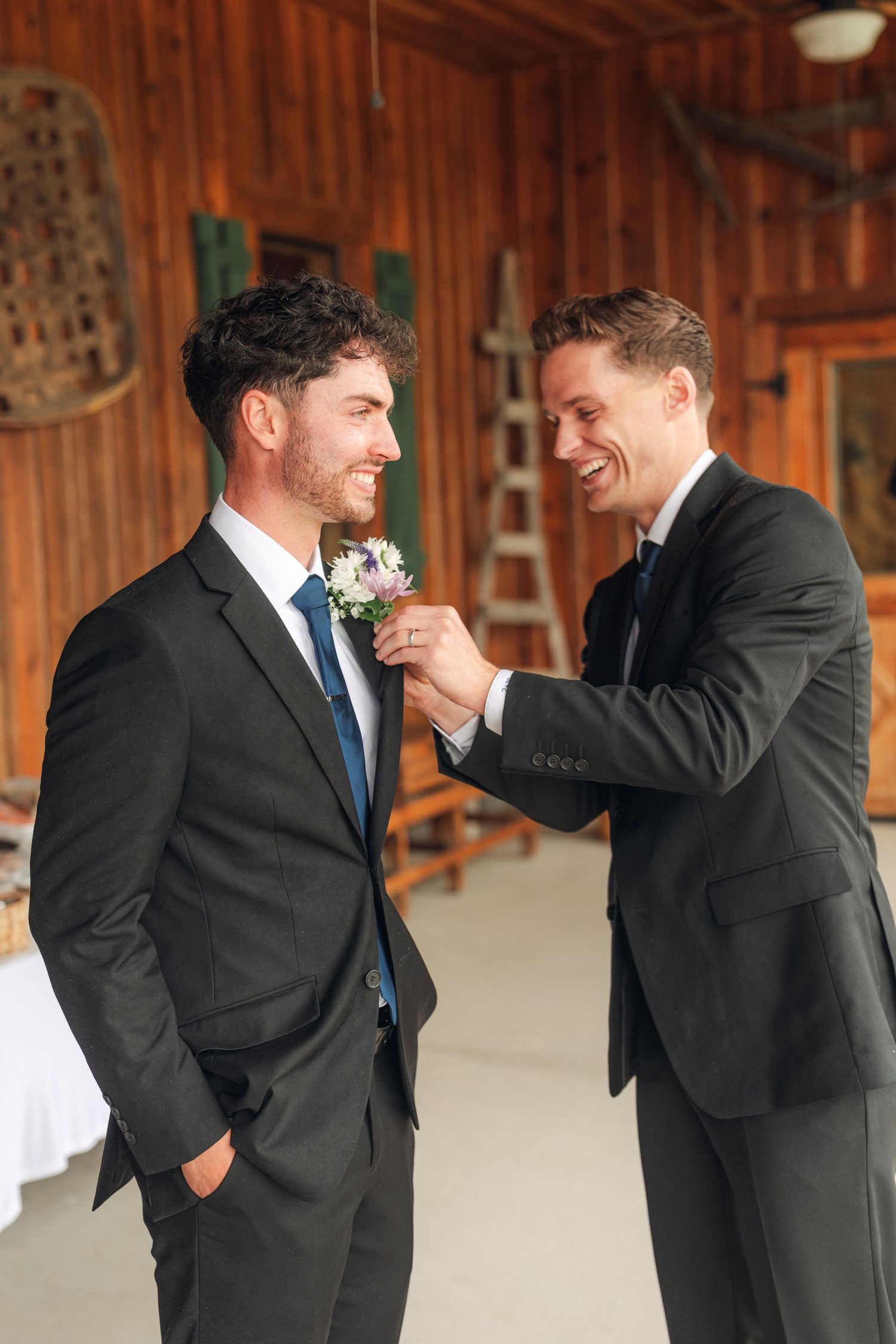 Two groomsmen in dark suits share a laugh while pinning a floral boutonniere inside a rustic wooden barn.