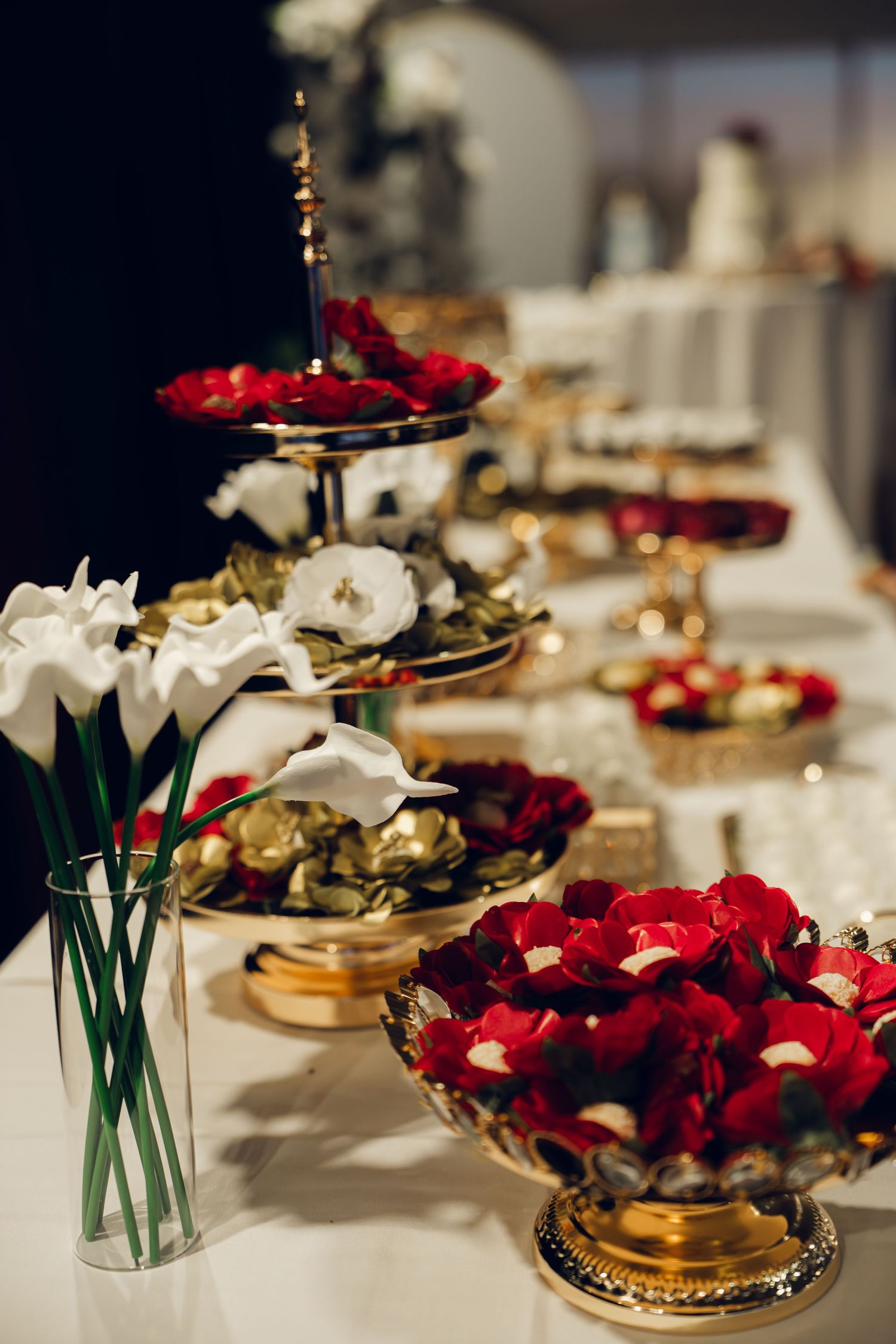 Luxurious wedding candy table with red rose petals in gold bowls and white calla lily arrangements.