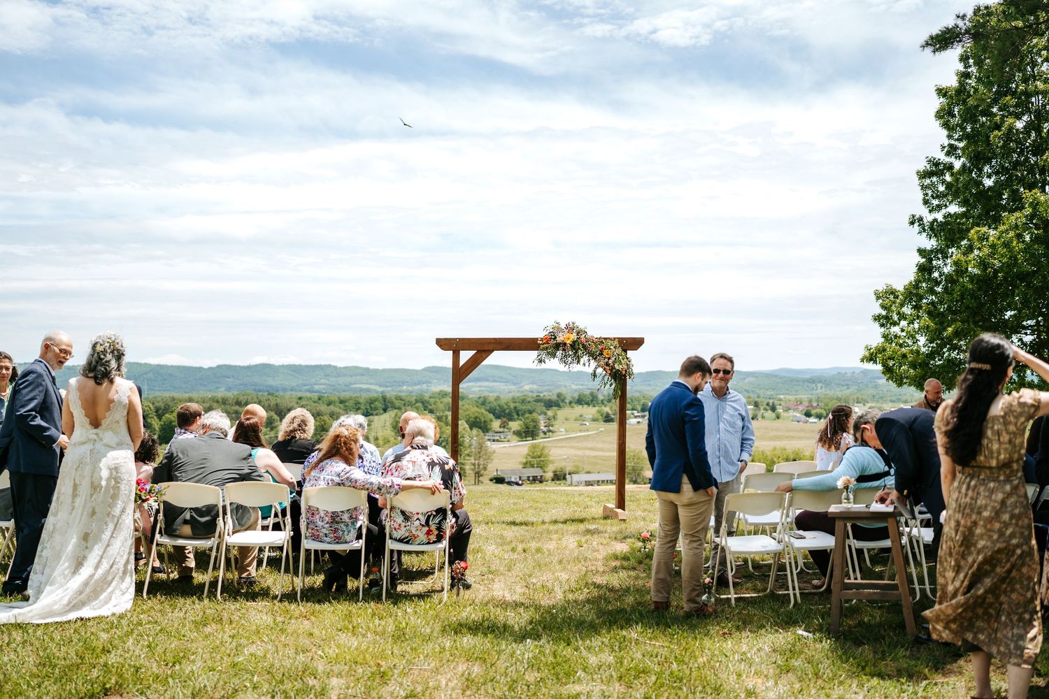 Outdoor wedding ceremony on a hilltop meadow with wooden arch and scenic countryside views.