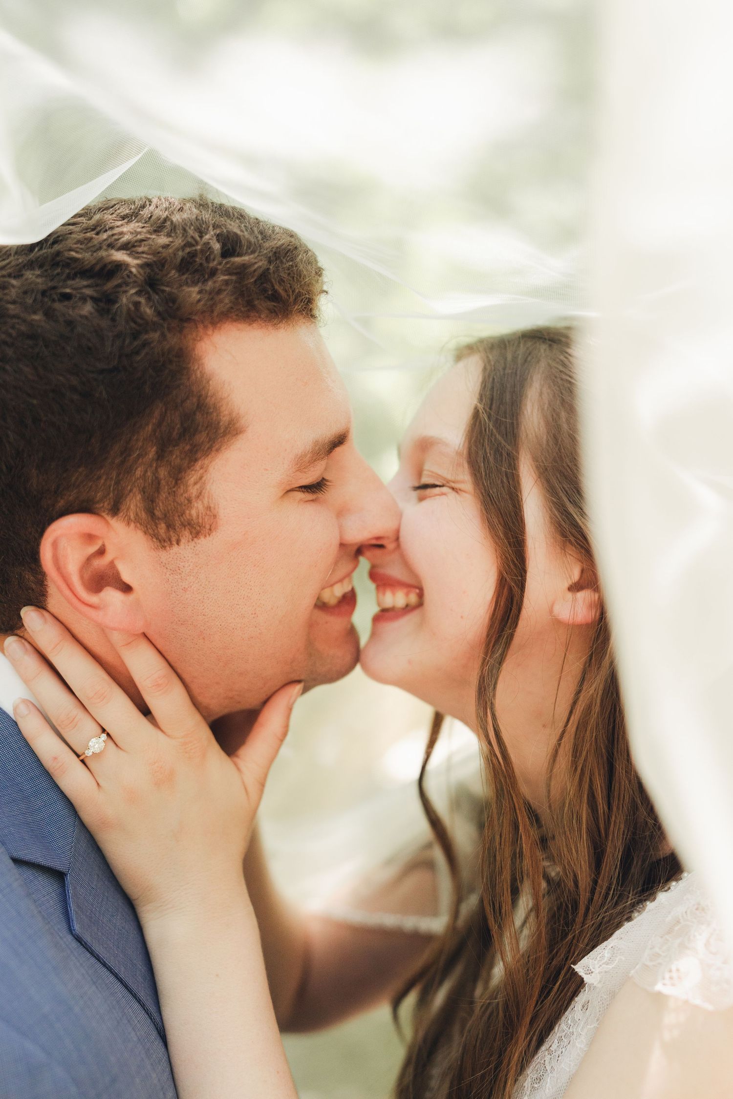 Bride and groom sharing a romantic almost-kiss under a white wedding veil, smiling joyfully on their wedding day.
