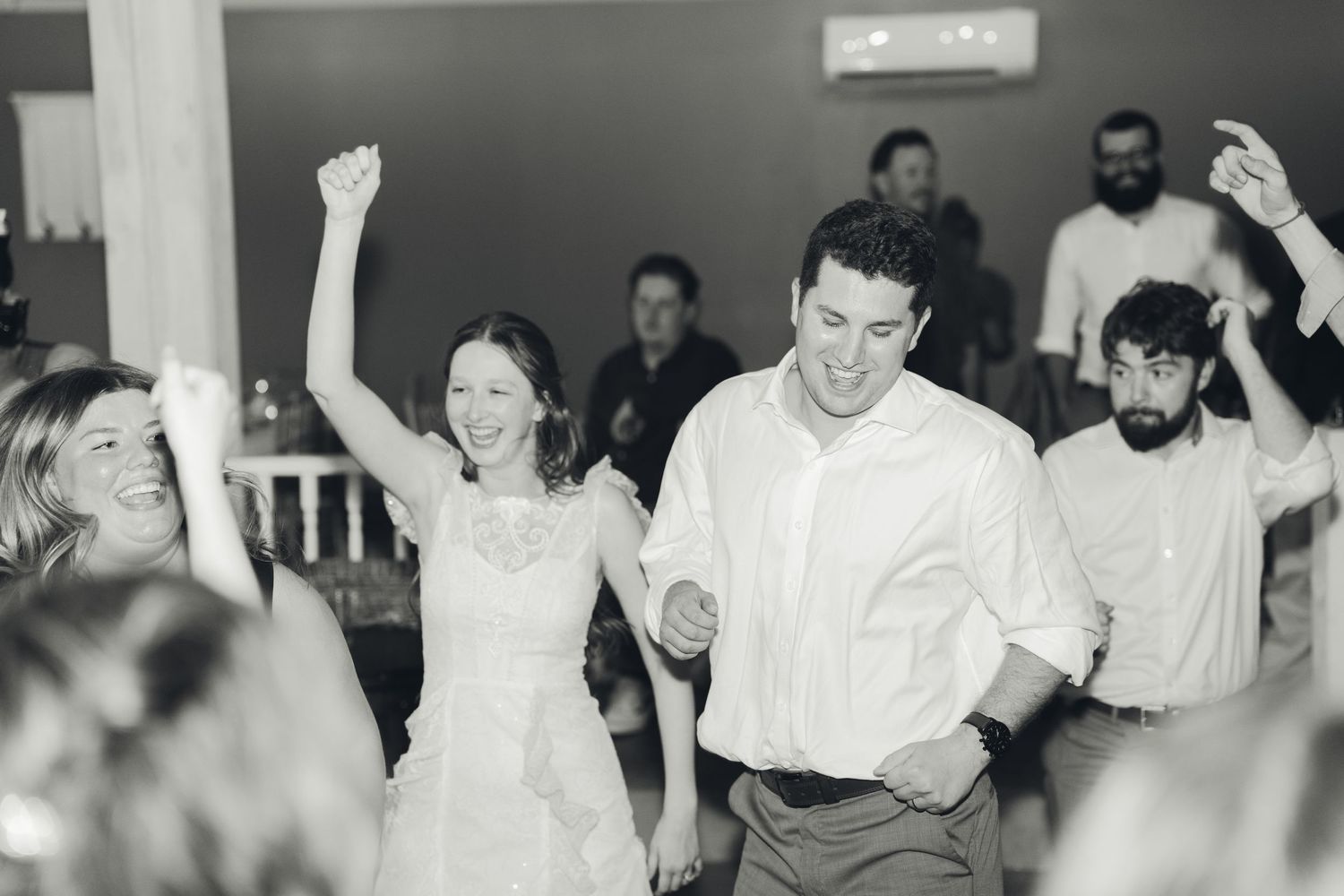 Black and white photo of bride and groom dancing joyfully with guests at wedding reception.