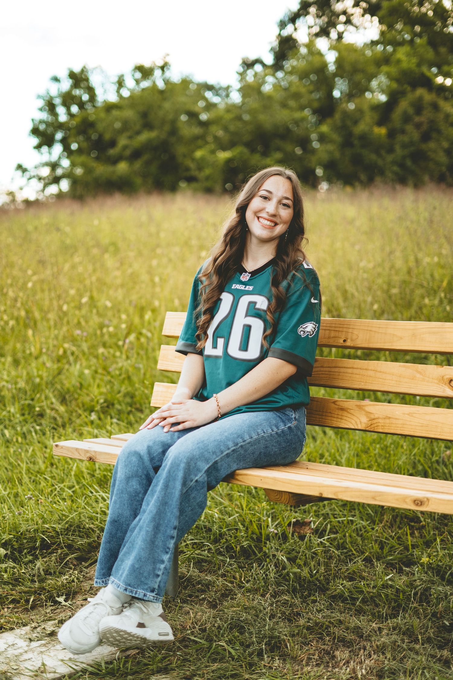 Smiling teen girl in Eagles jersey number 26 sits on wooden bench in green field.