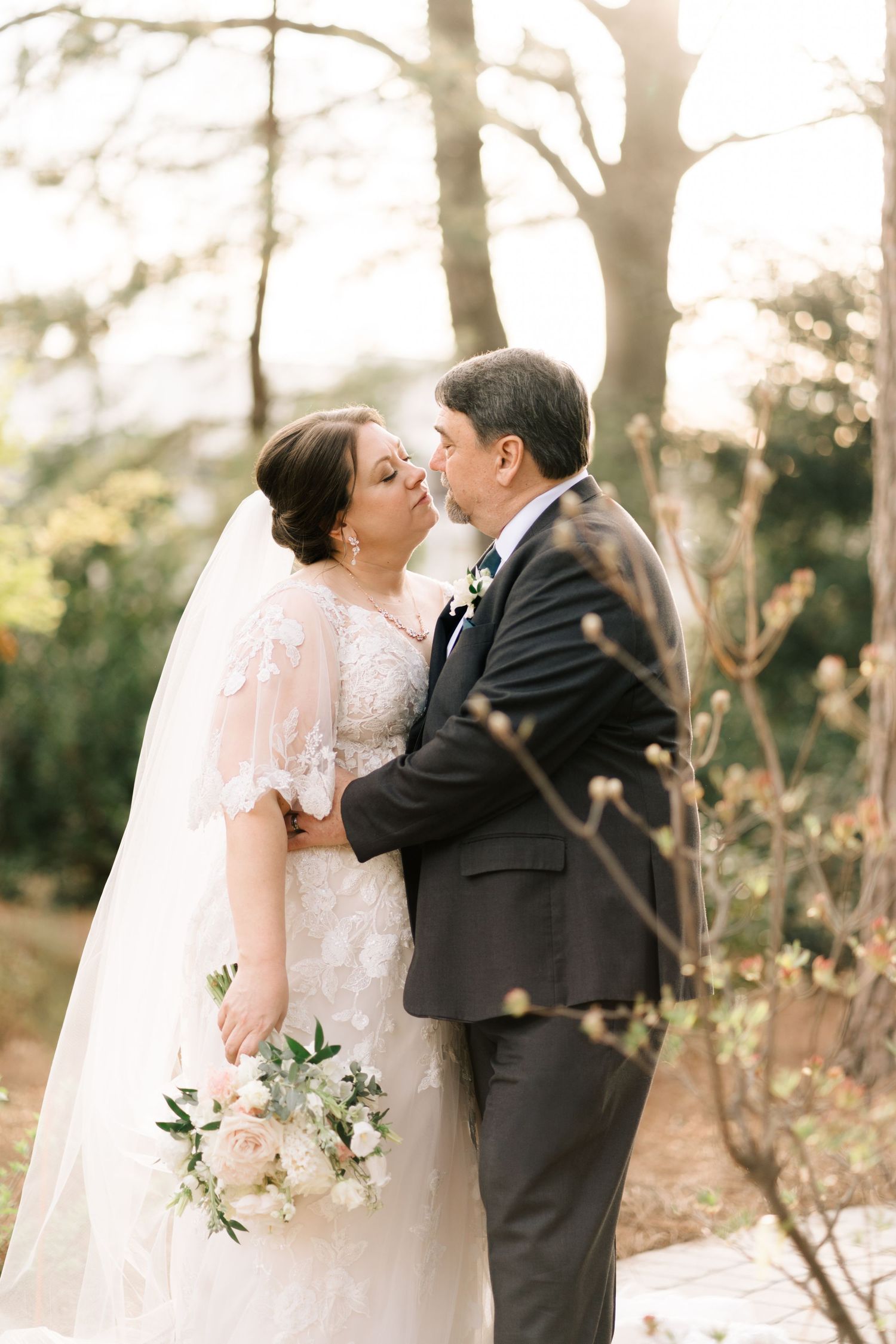 Bride and groom leaning in for a kiss outdoors, surrounded by soft natural light and greenery.