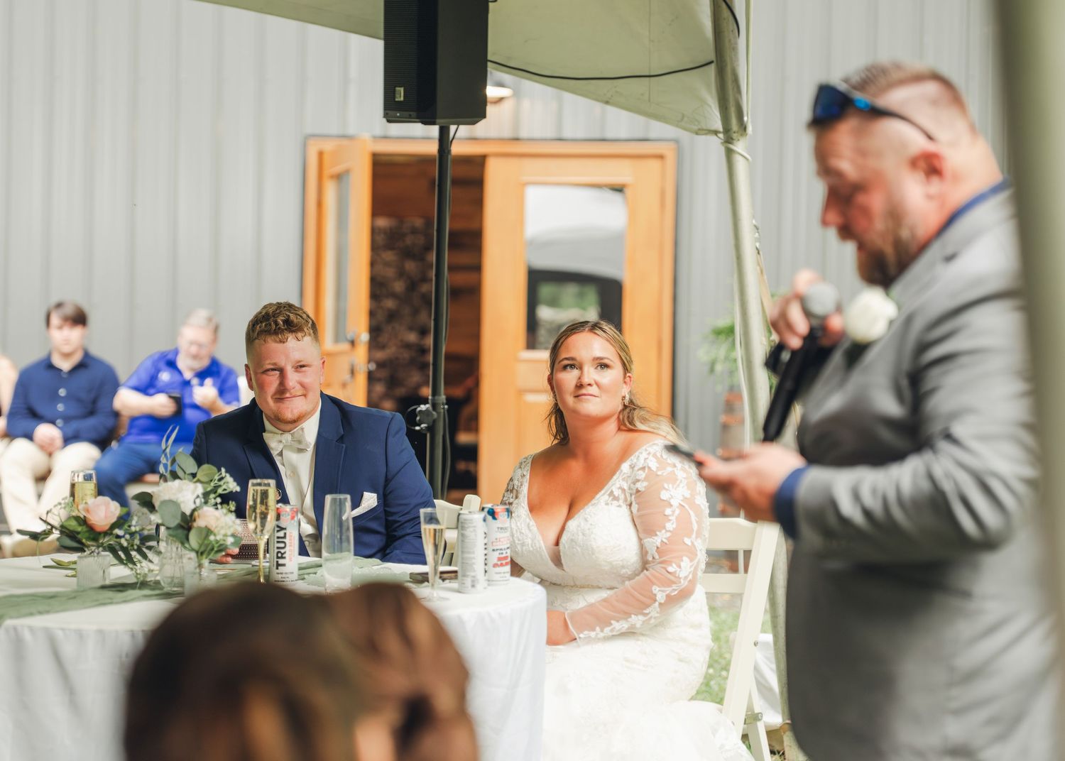 Bride and groom listen to best man give speech at elegant outdoor wedding reception under tent.
