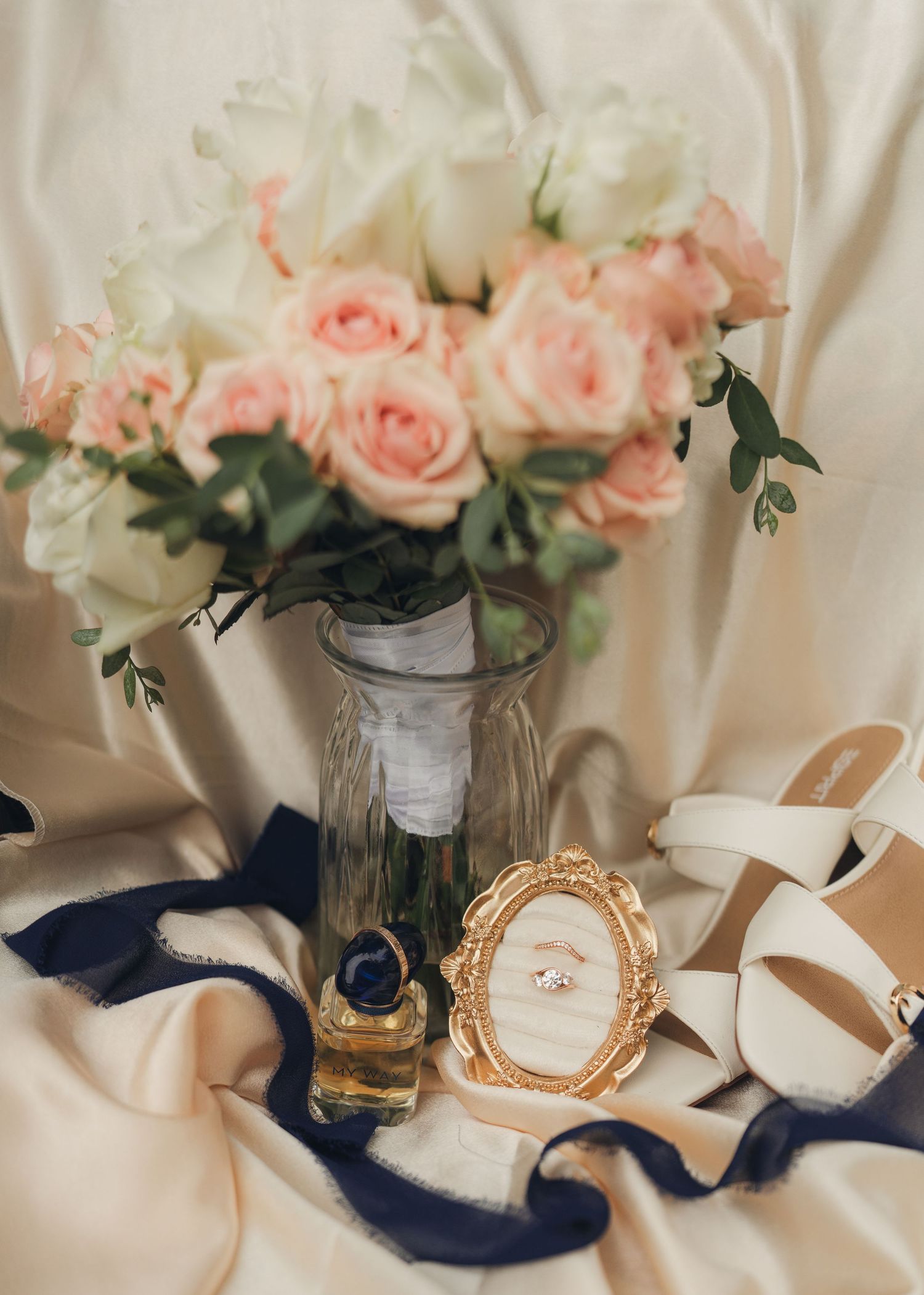 Elegant wedding flat lay with pink roses bouquet, perfume bottle, gold ring frame, and white heels on satin fabric.