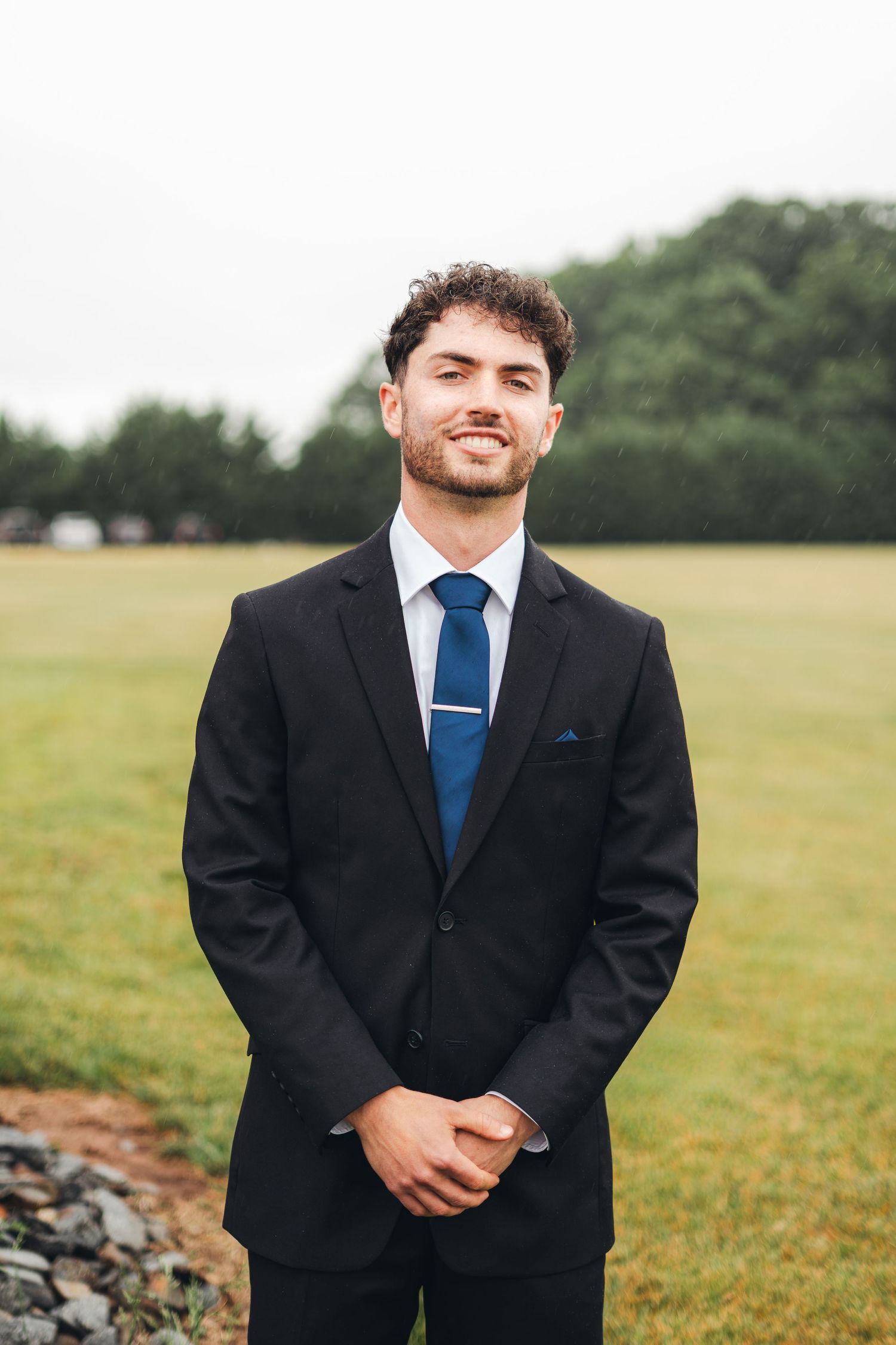 A young man in a black suit and blue tie smiles outdoors at a wedding ceremony on a cloudy day.