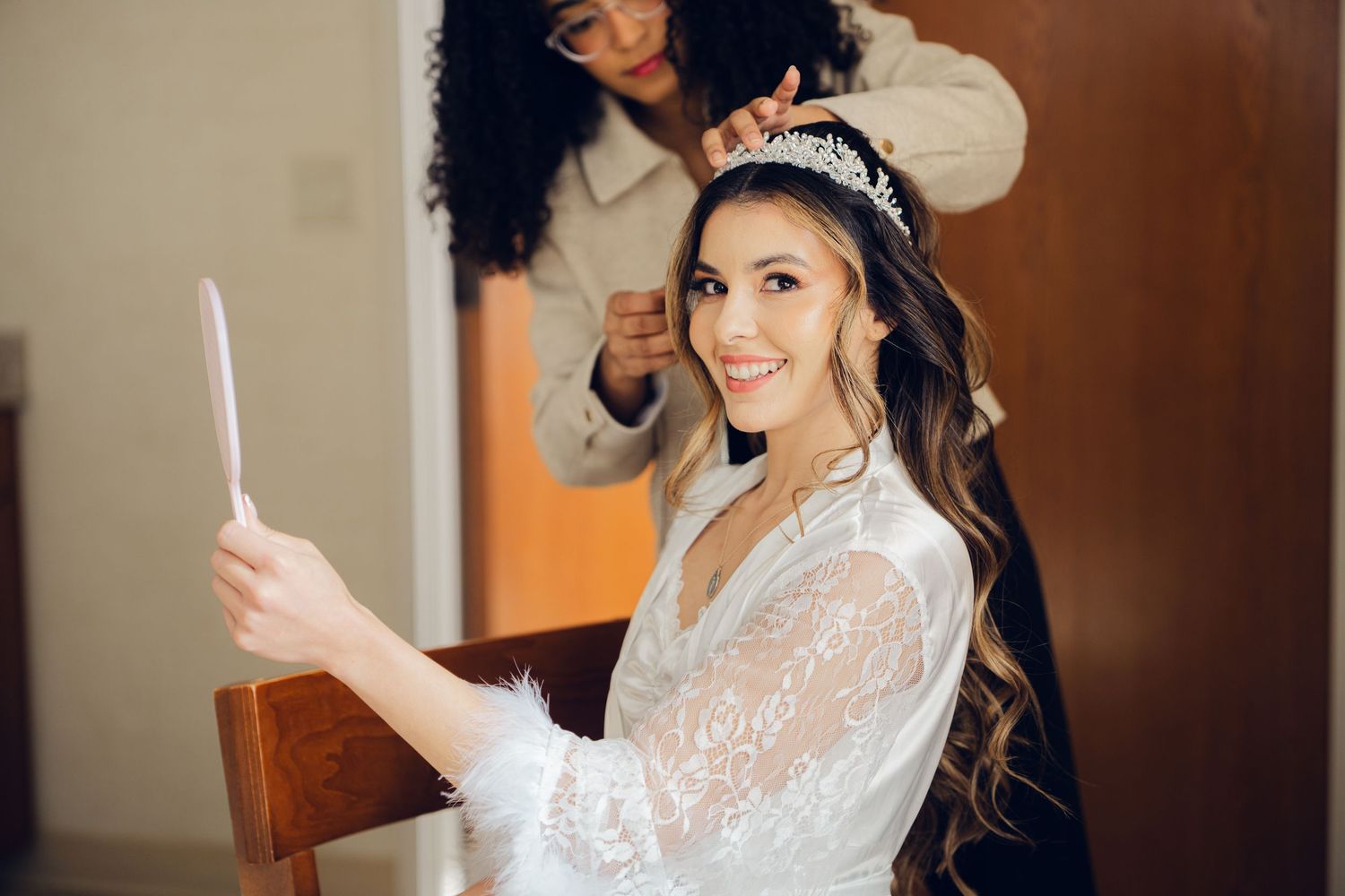 Smiling bride in lace robe holding mirror while hairstylist places tiara on her hair.