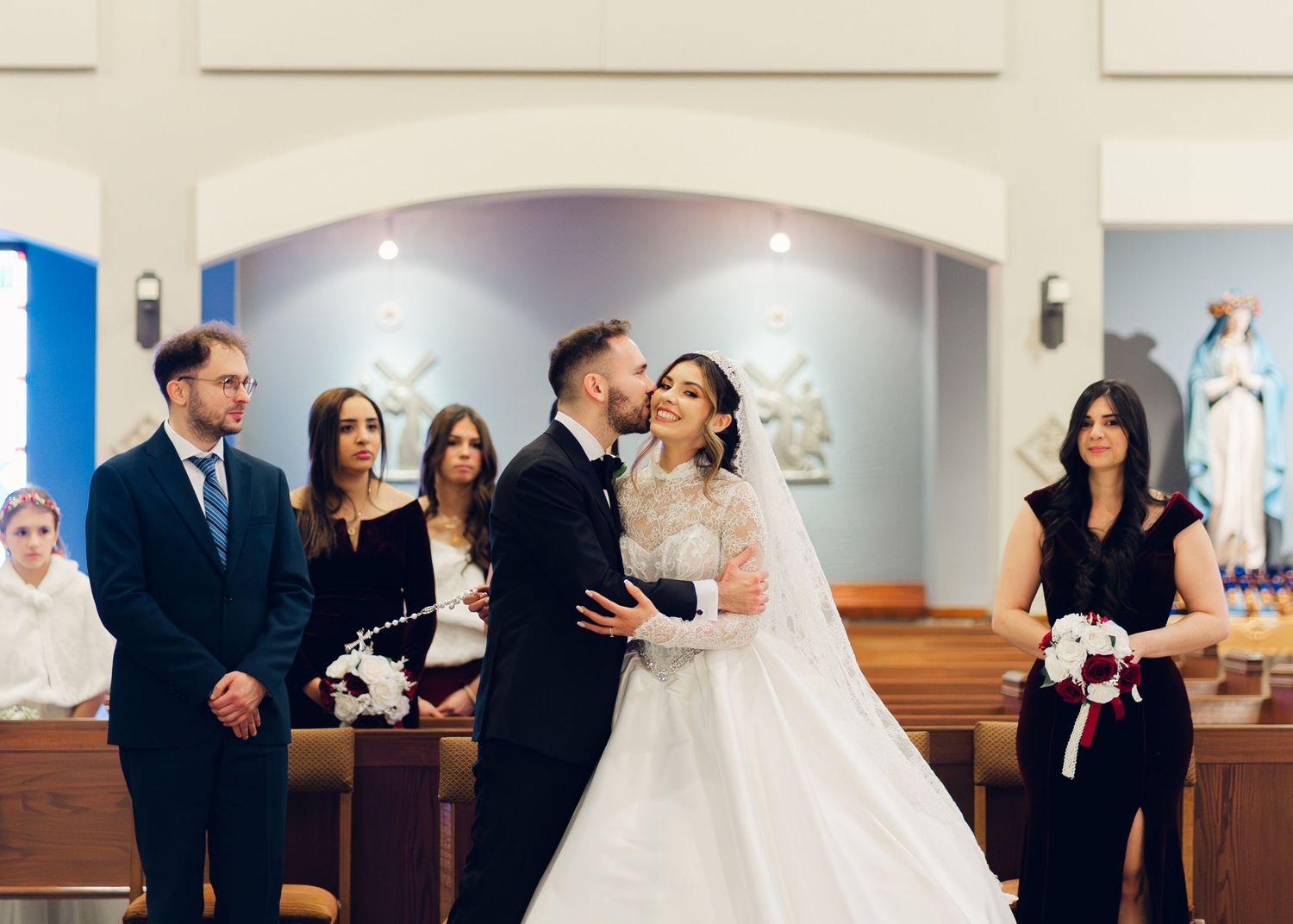 Bride and groom share first kiss at church altar surrounded by wedding party during ceremony.