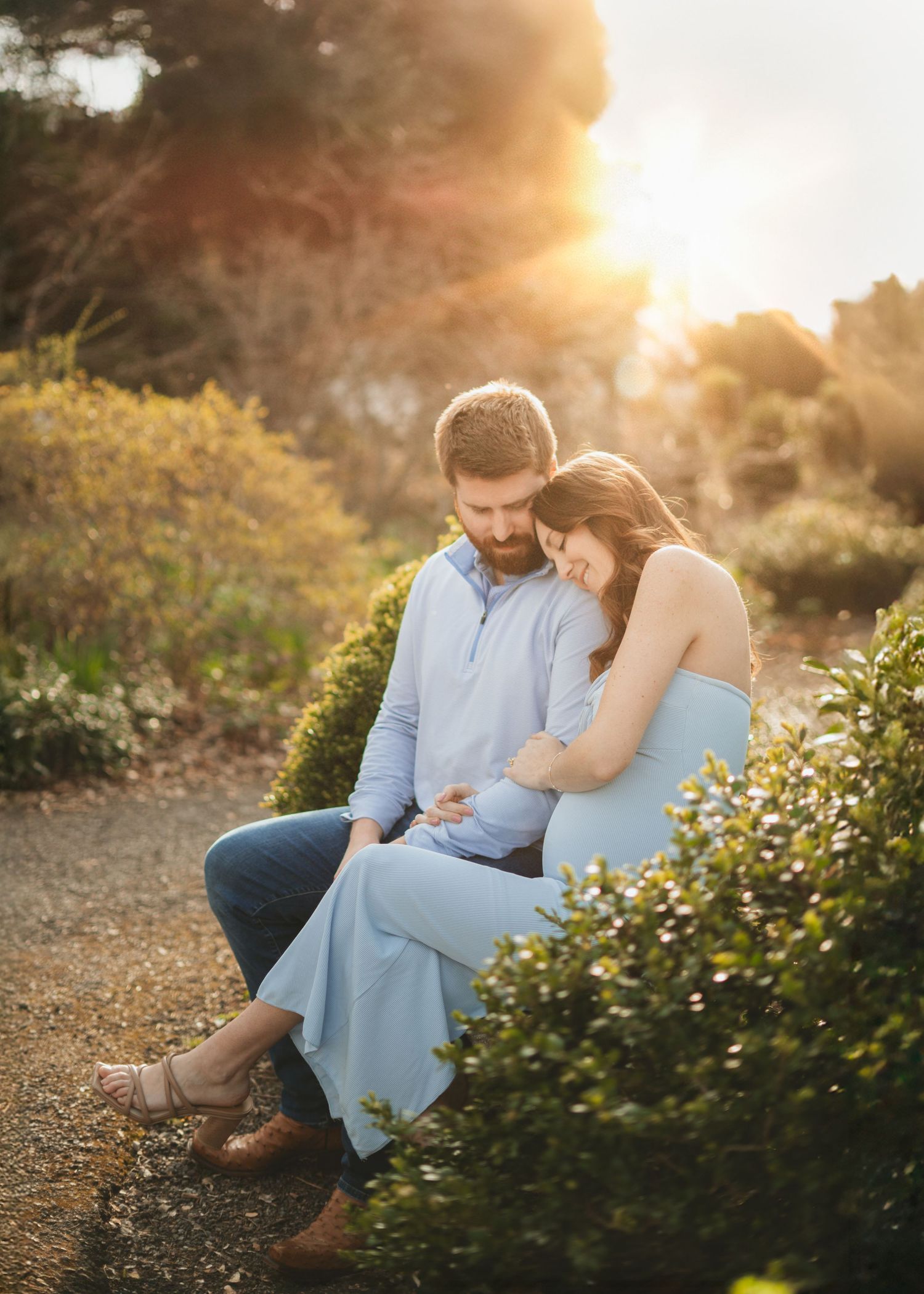 Couple sitting together at golden sunset, woman in blue dress leaning on mans shoulder outdoors.