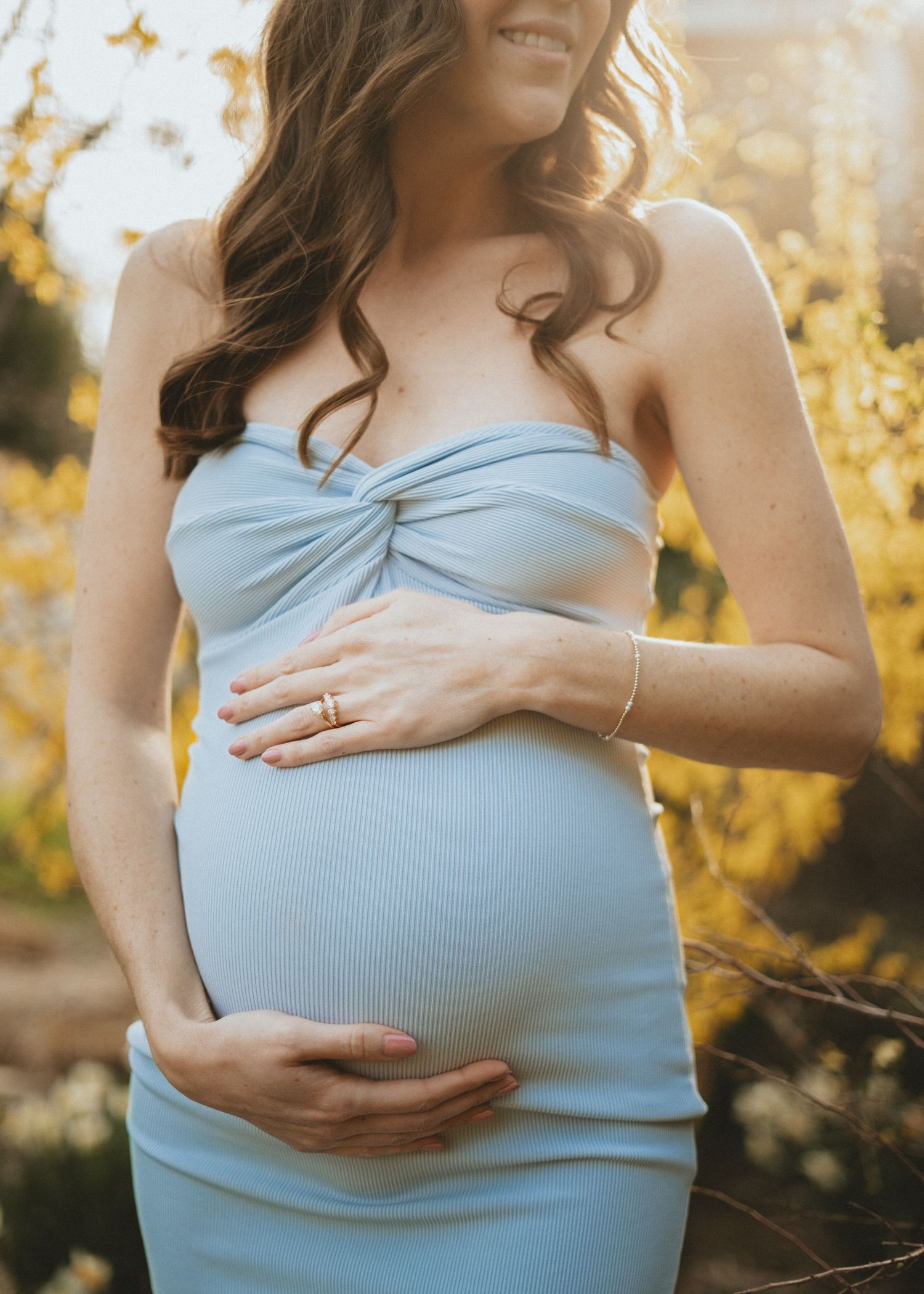 Pregnant woman in a light blue strapless dress cradling her baby bump during a glowing outdoor maternity shoot.