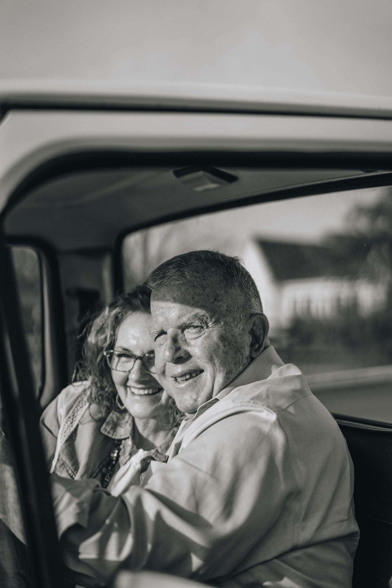 Black and white photo of smiling older couple embracing inside vintage pickup truck cab.