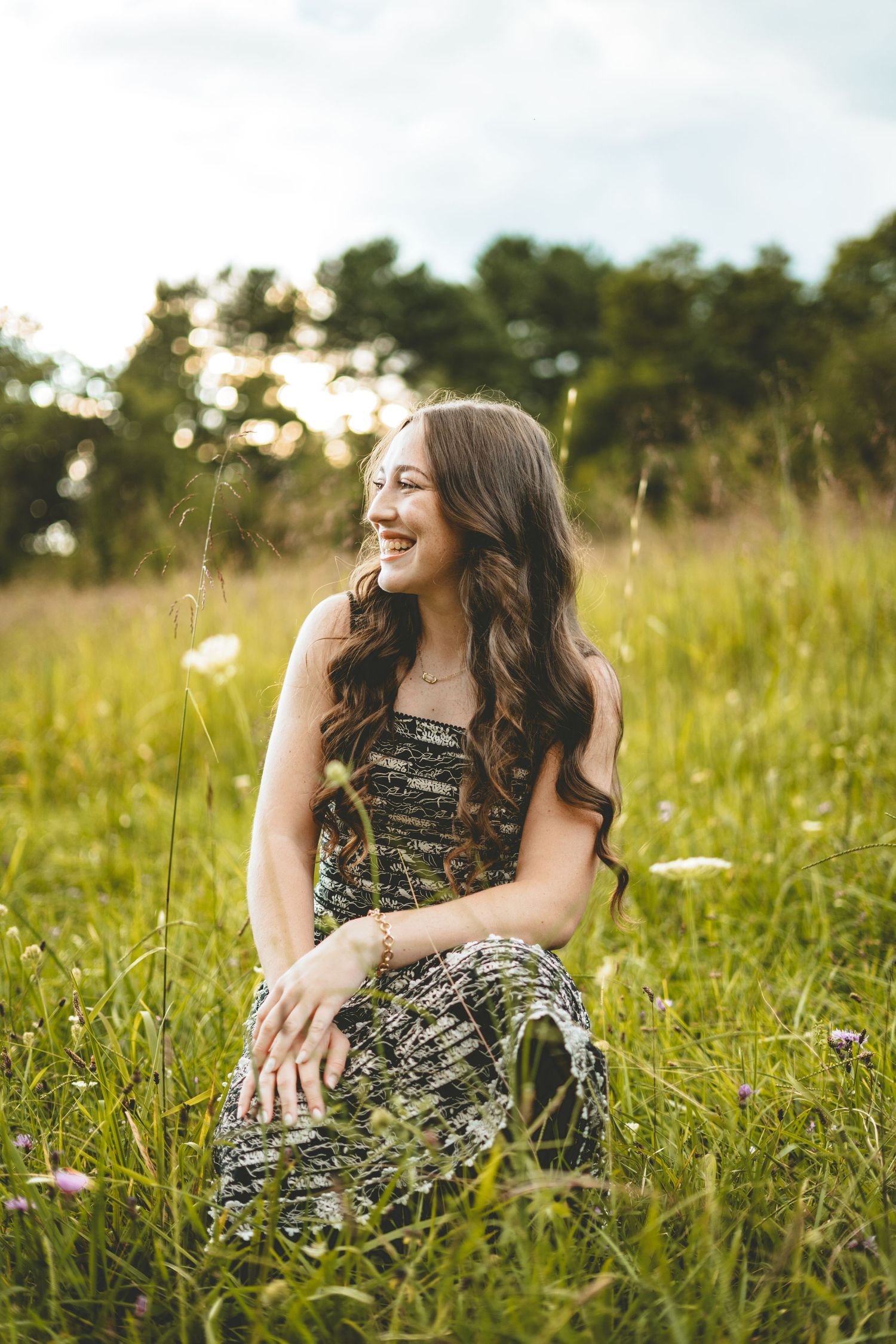 Young woman with long curly hair sitting in sunny green meadow laughing.