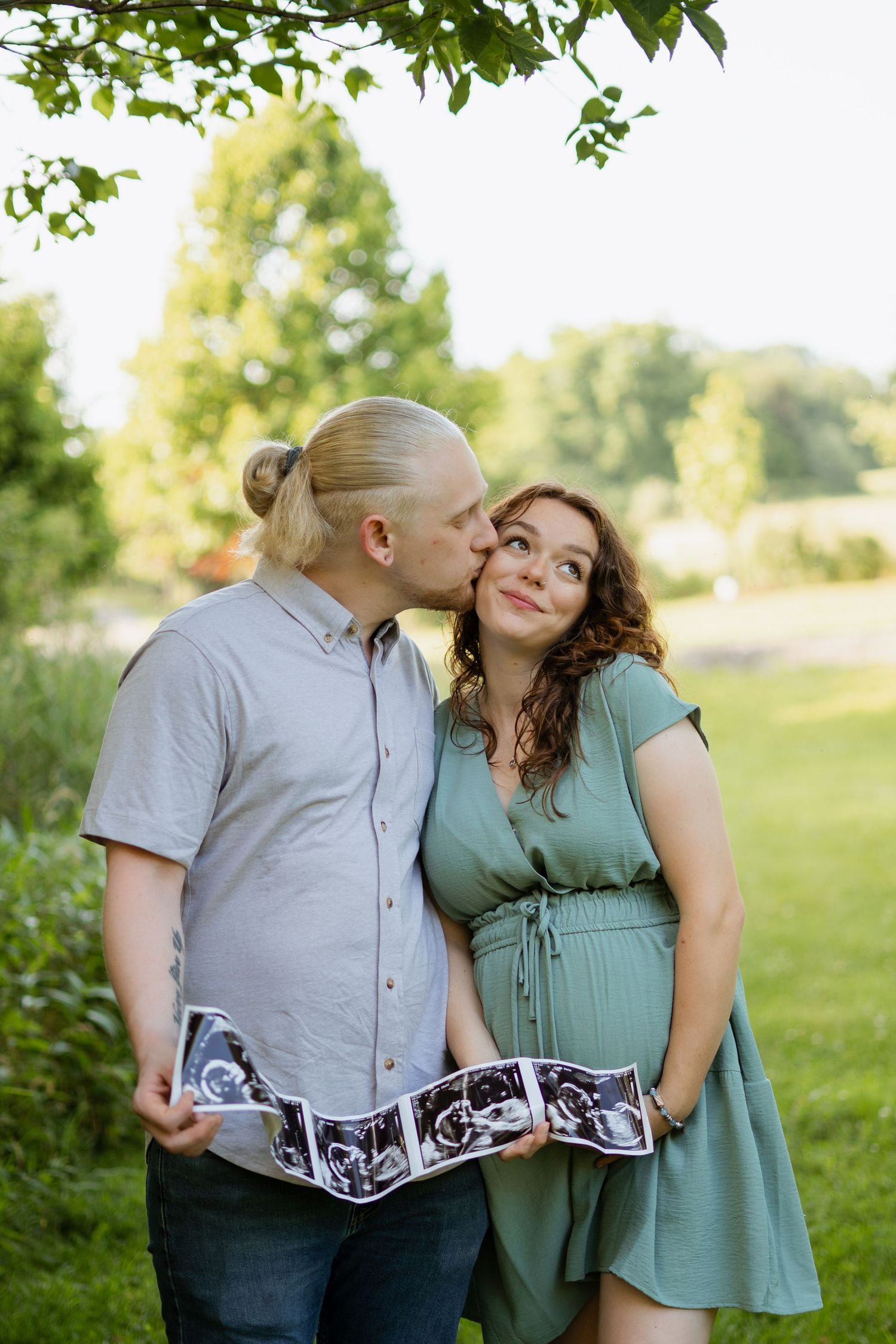 Expecting couple holding ultrasound photos outdoors, man kissing woman's cheek in a lush green park setting.