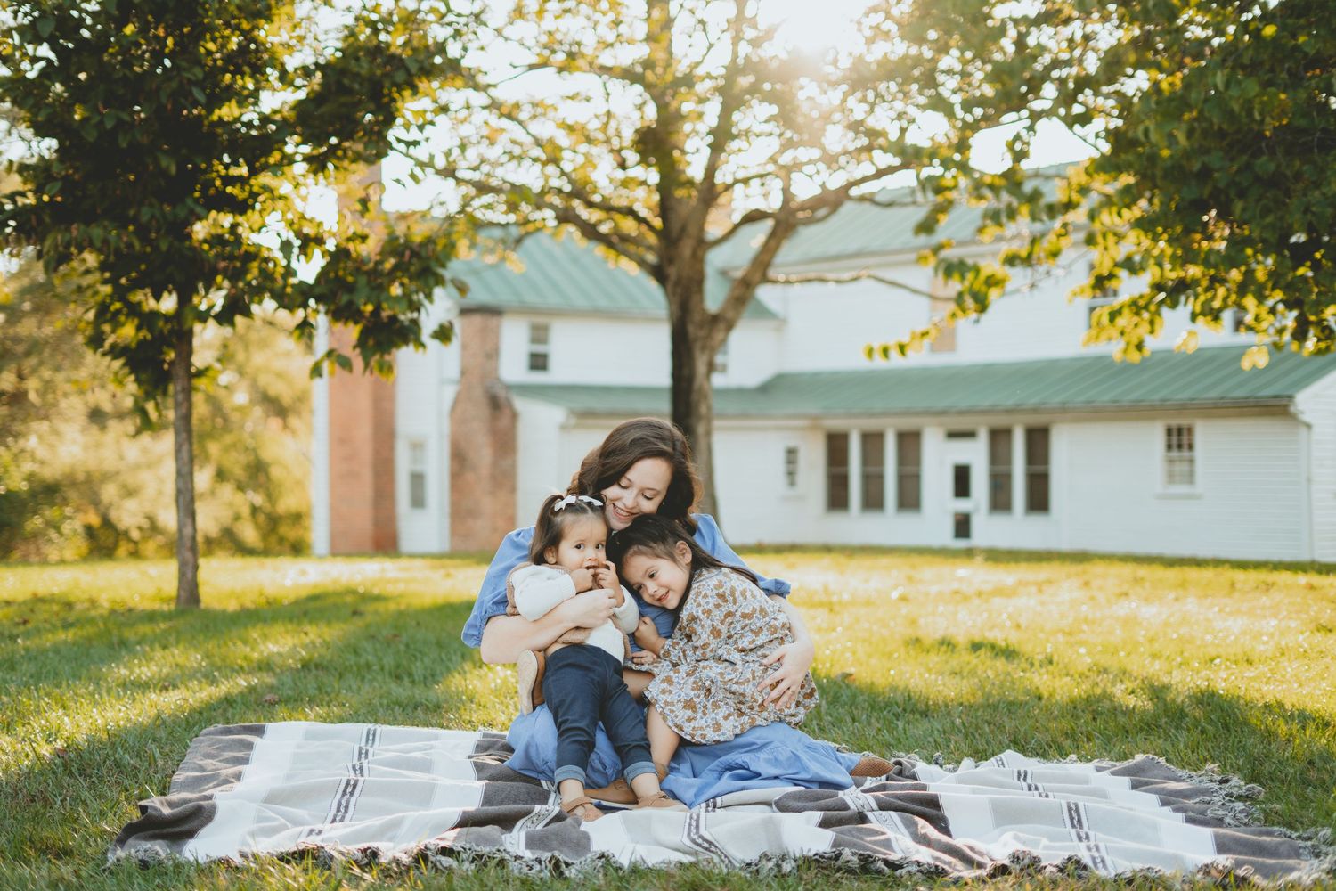 Mother sitting on blanket in sunny yard cuddling two young daughters near white farmhouse.