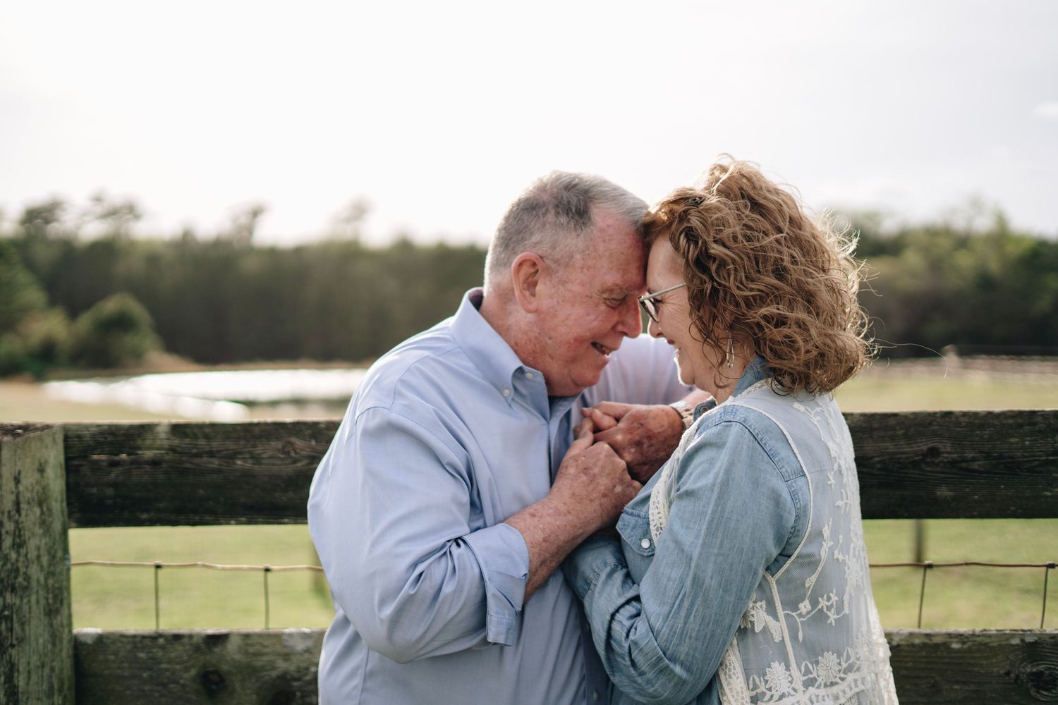 Romantic older couple holding hands and touching foreheads tenderly near wooden fence on farm.