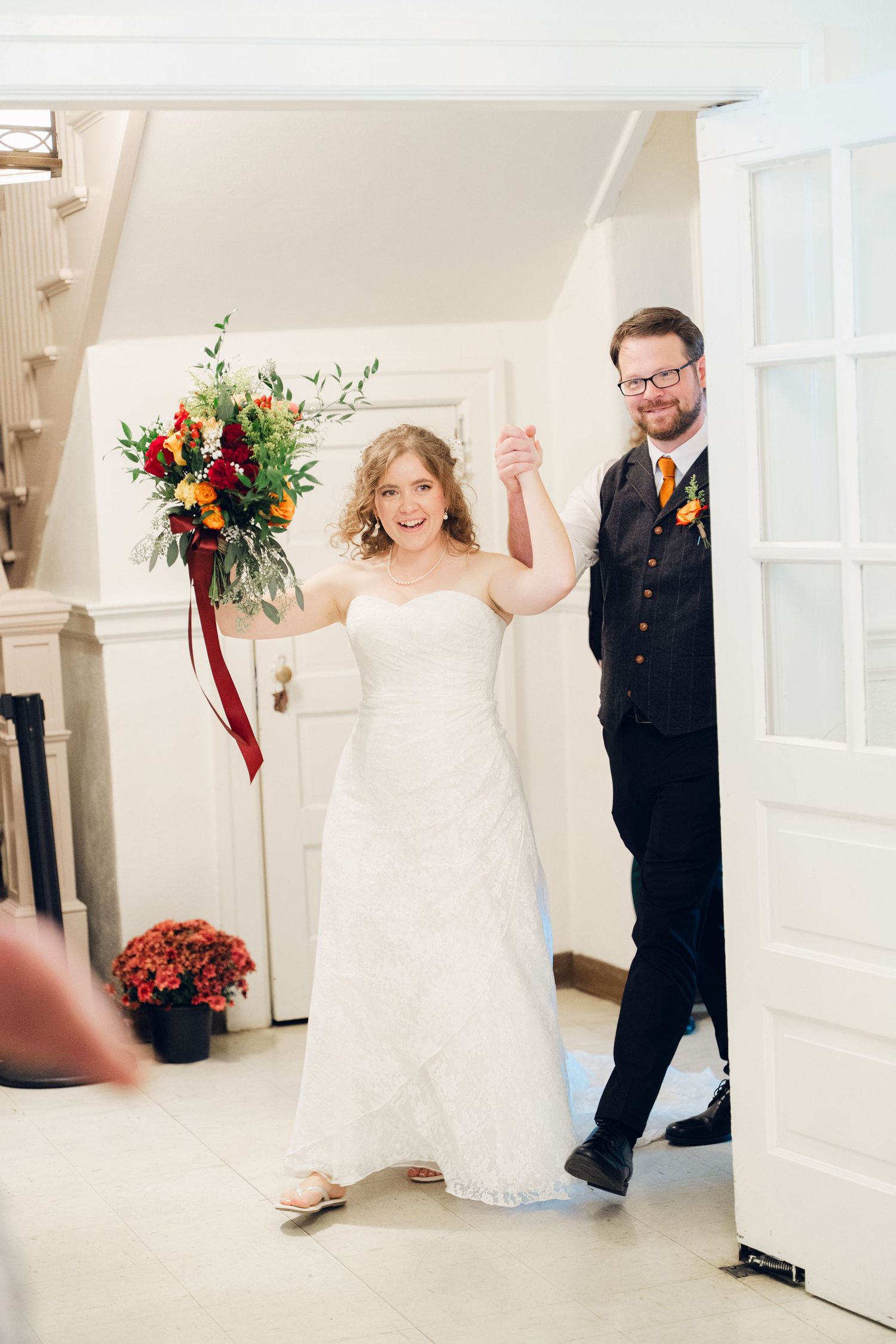 Joyful bride and groom make a grand entrance holding hands and raising a fall bouquet.