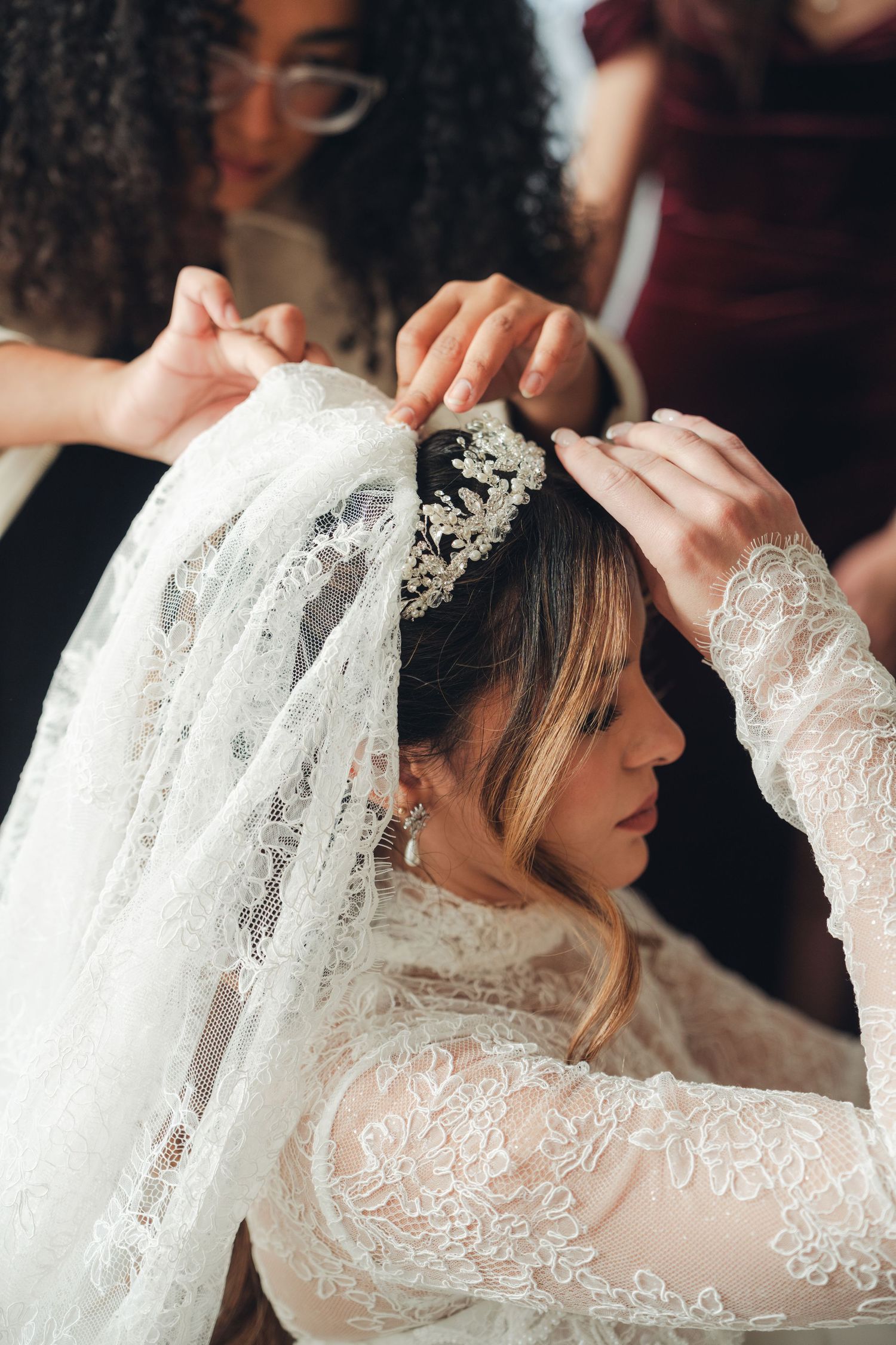 Bridesmaids placing crystal tiara on bride wearing lace wedding dress and veil.
