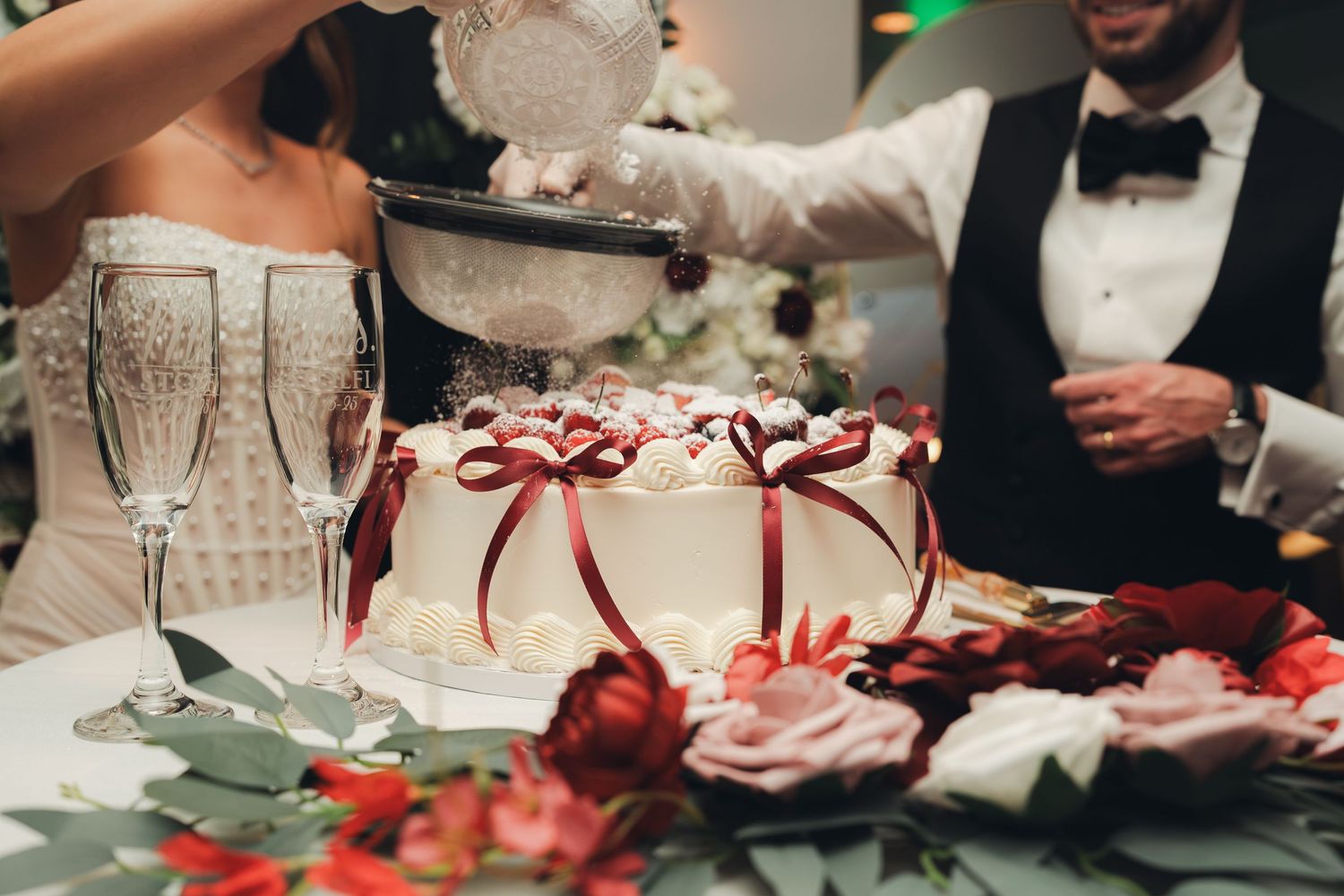 Bride and groom cutting elegant white wedding cake adorned with red ribbon bows and roses.
