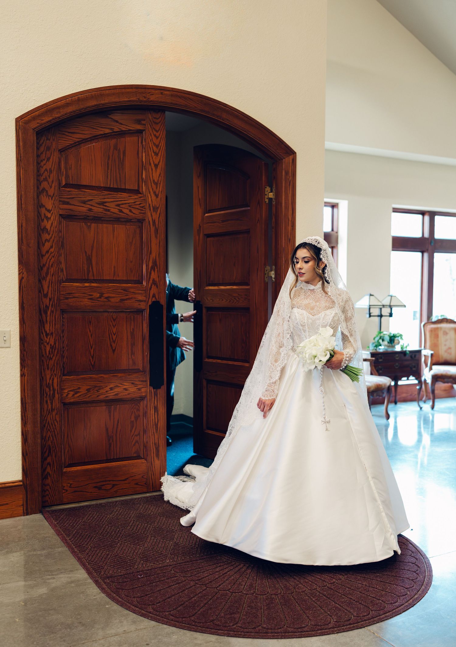 Elegant bride in lace ball gown and veil standing before arched wooden doors holding bouquet.
