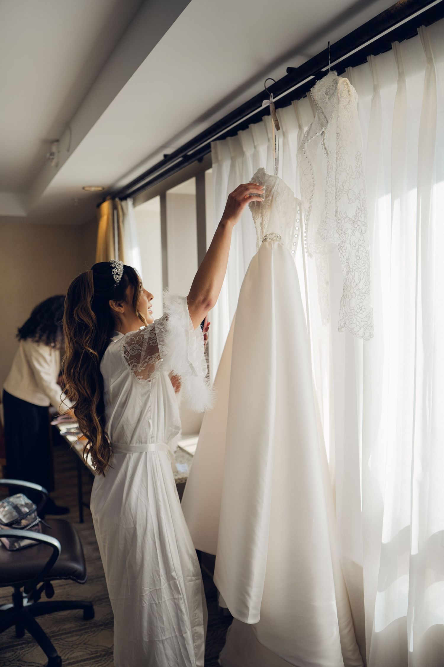 A bride in a white lace robe reaches for her wedding dress hanging by a sunlit window.