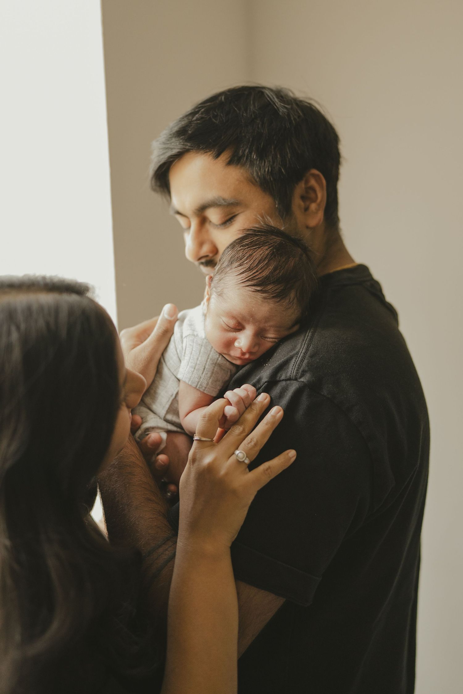 Father tenderly holding sleeping newborn while mother gently touches baby in soft indoor light.