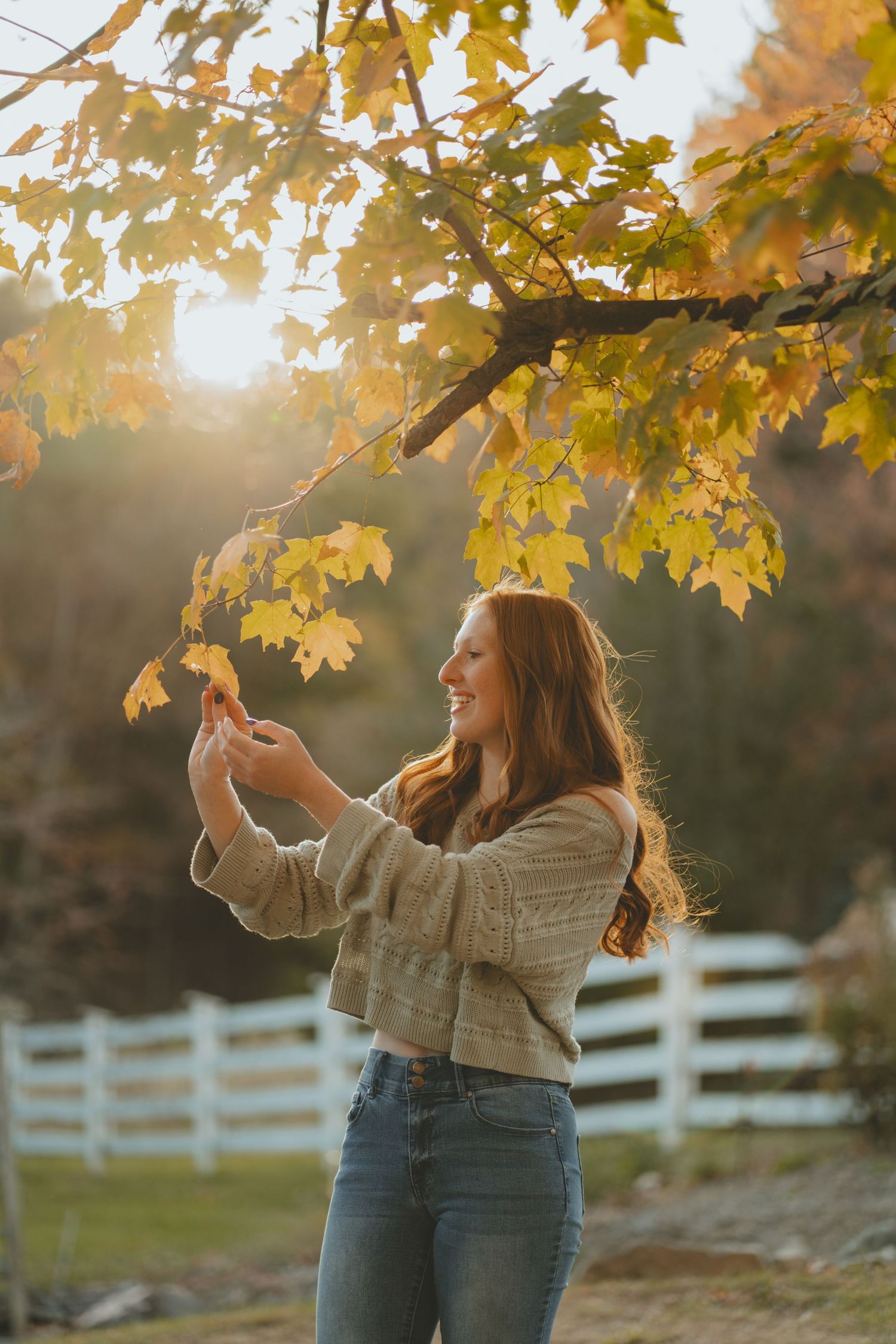 Smiling redhead girl touching yellow maple leaves in golden autumn sunlight.