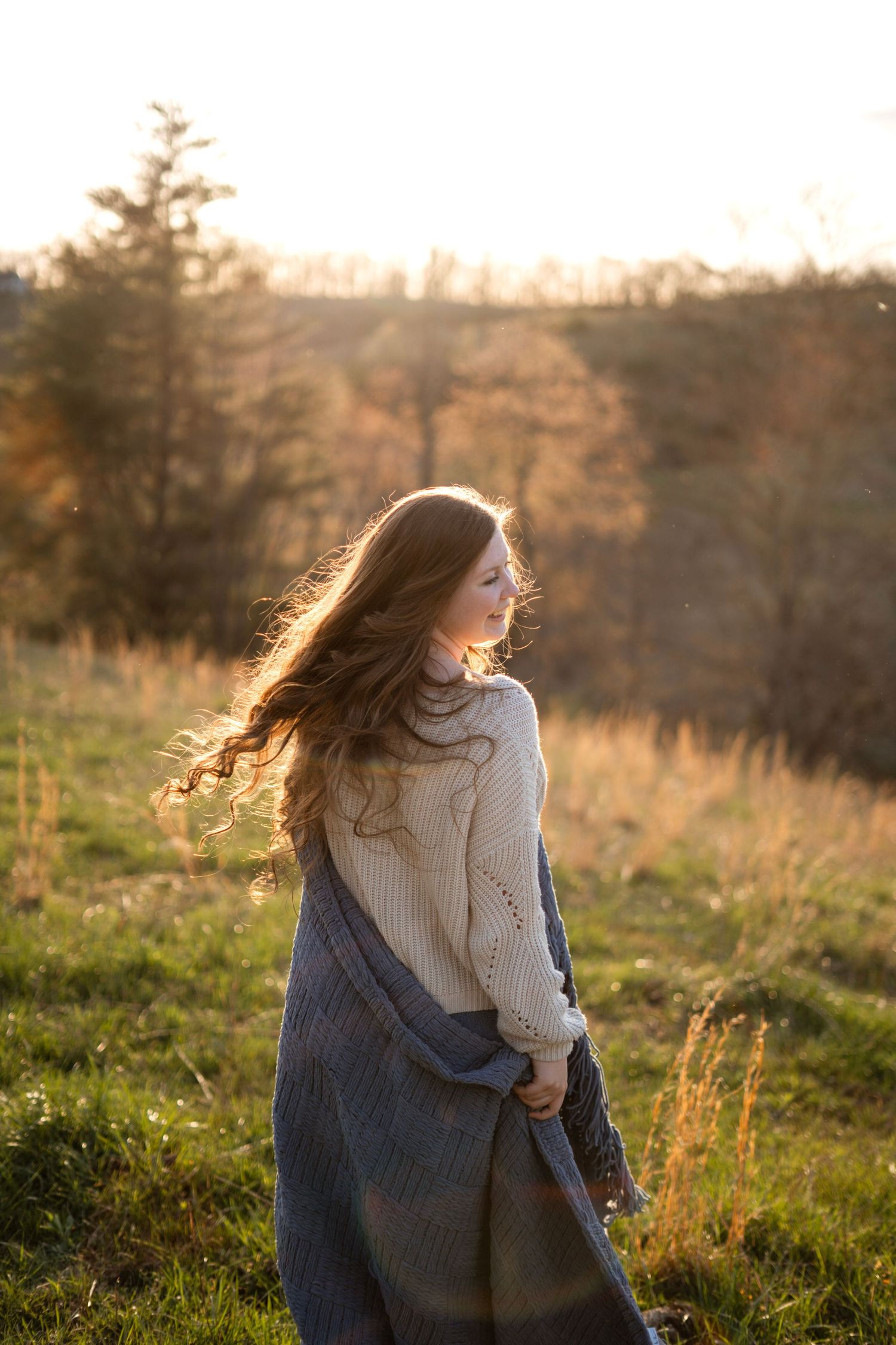 Young woman with long auburn hair in cream sweater and plaid wrap standing in golden sunset field.
