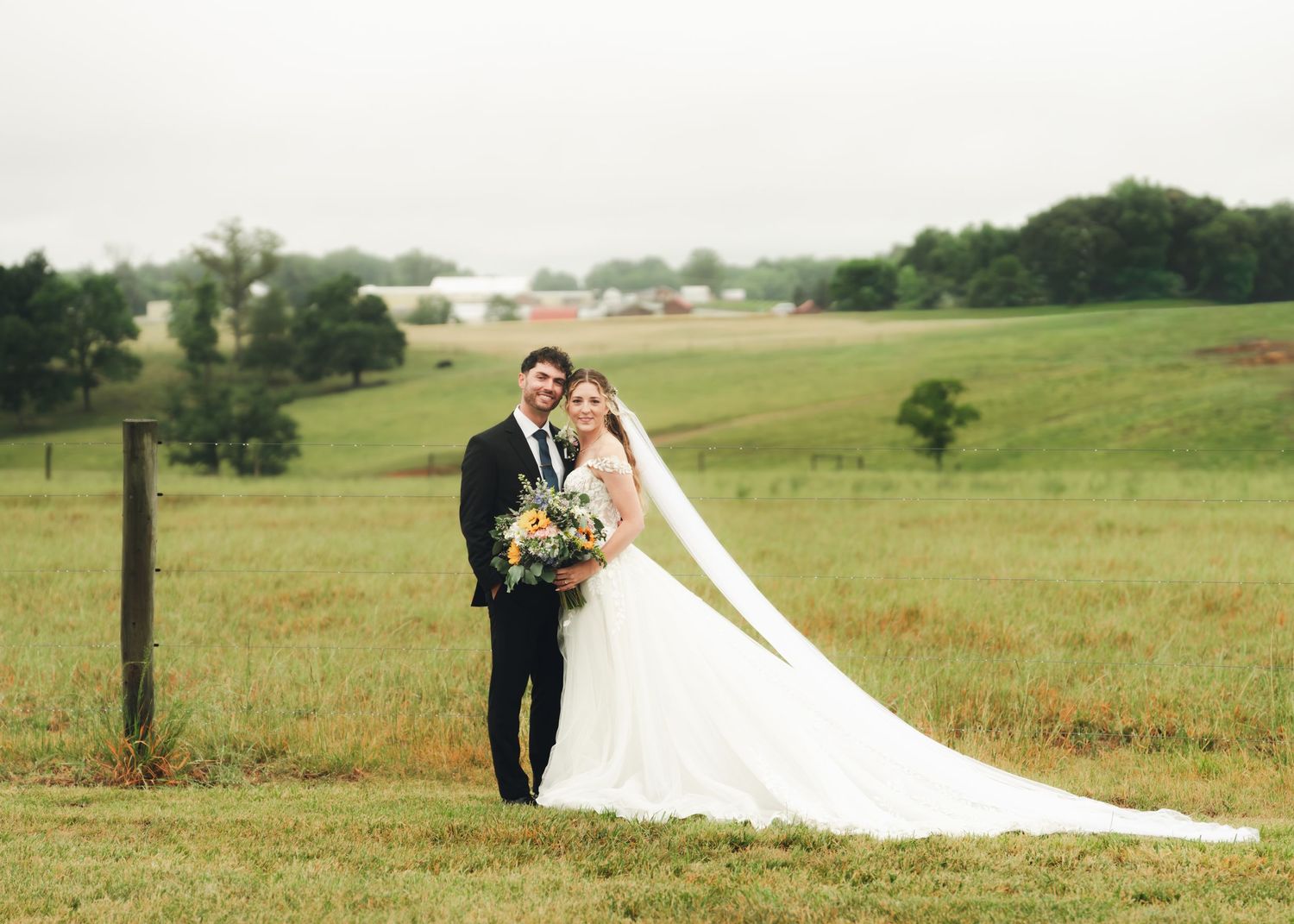 Bride and groom pose romantically in a lush green countryside field, her long veil flowing in the breeze.