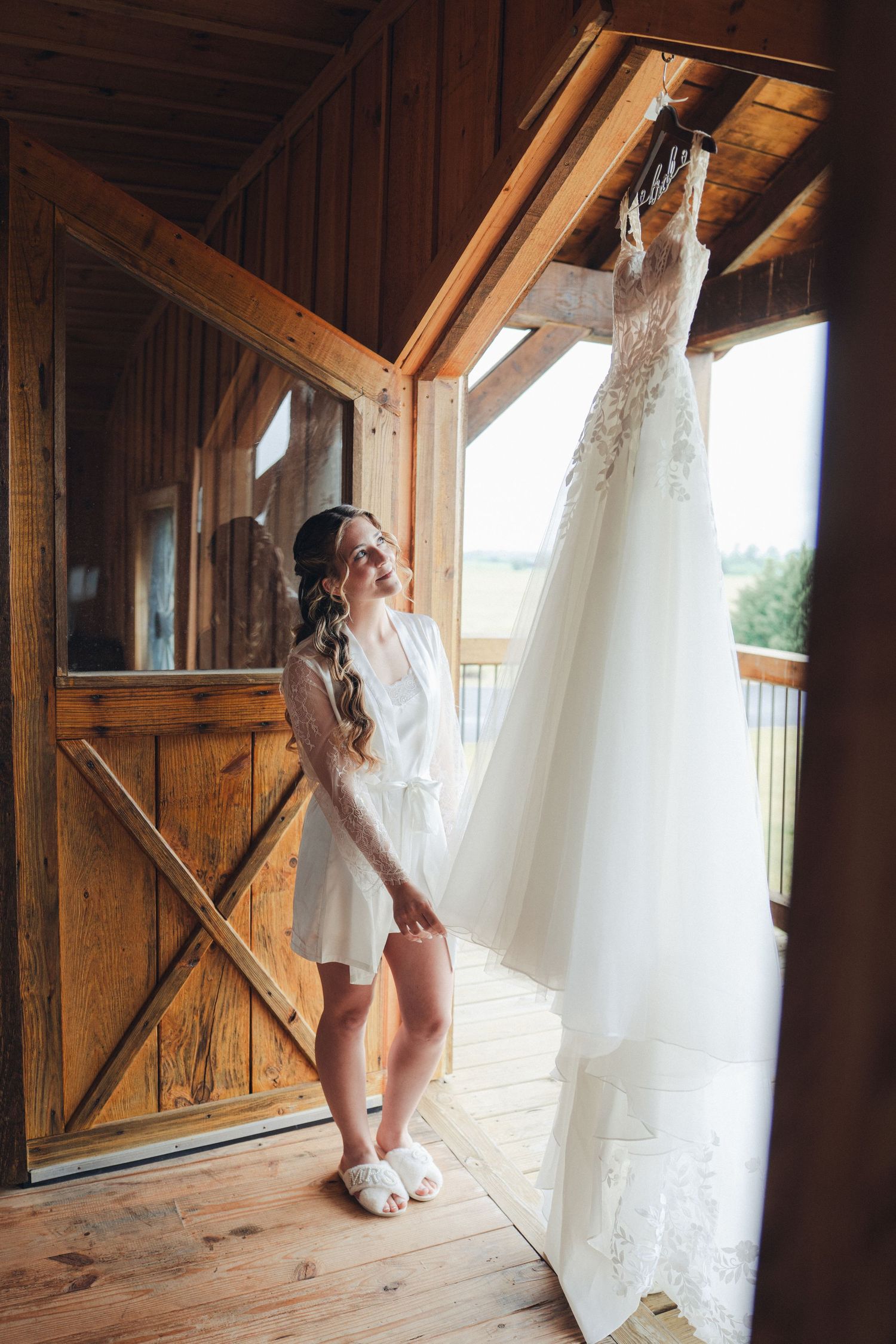 Bride in white robe reaching for her lace wedding dress hanging in a rustic wooden barn.