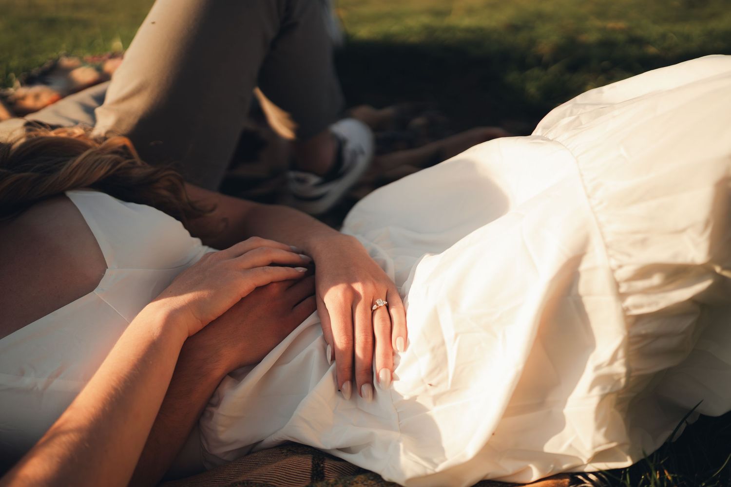 Close-up of couple holding hands in laps during outdoor golden hour engagement session, woman wearing white dress.