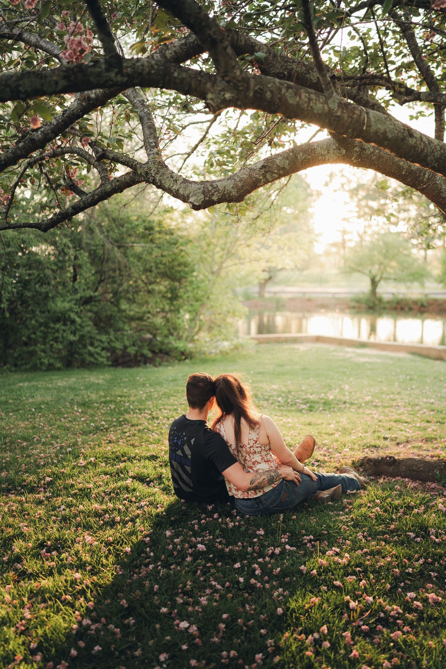 Couple sitting together on grass under blooming tree by a pond in golden hour light.