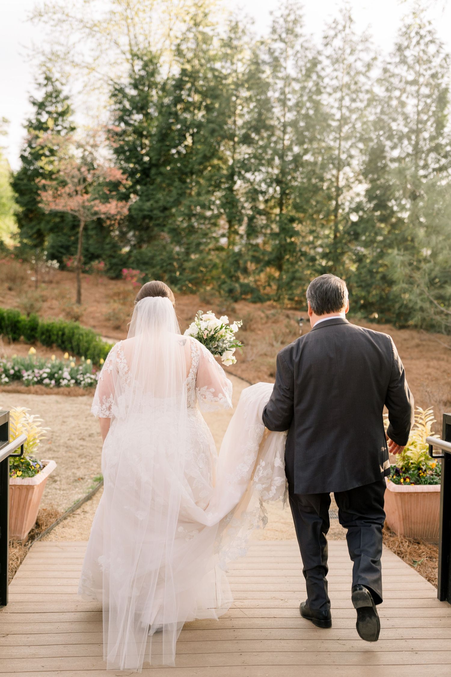 Bride in lace gown with veil walks arm-in-arm with escort along wooden path surrounded by lush trees.
