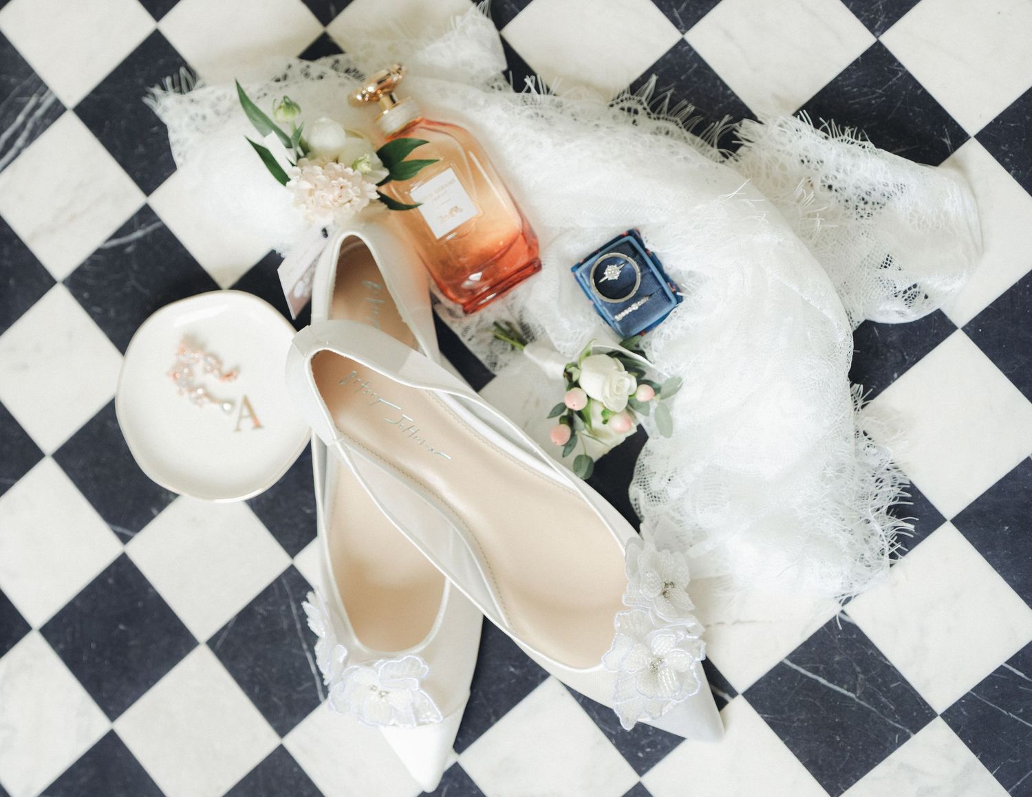 Elegant wedding flat lay featuring white bridal heels, perfume, ring box, and floral accents on checkered floor.