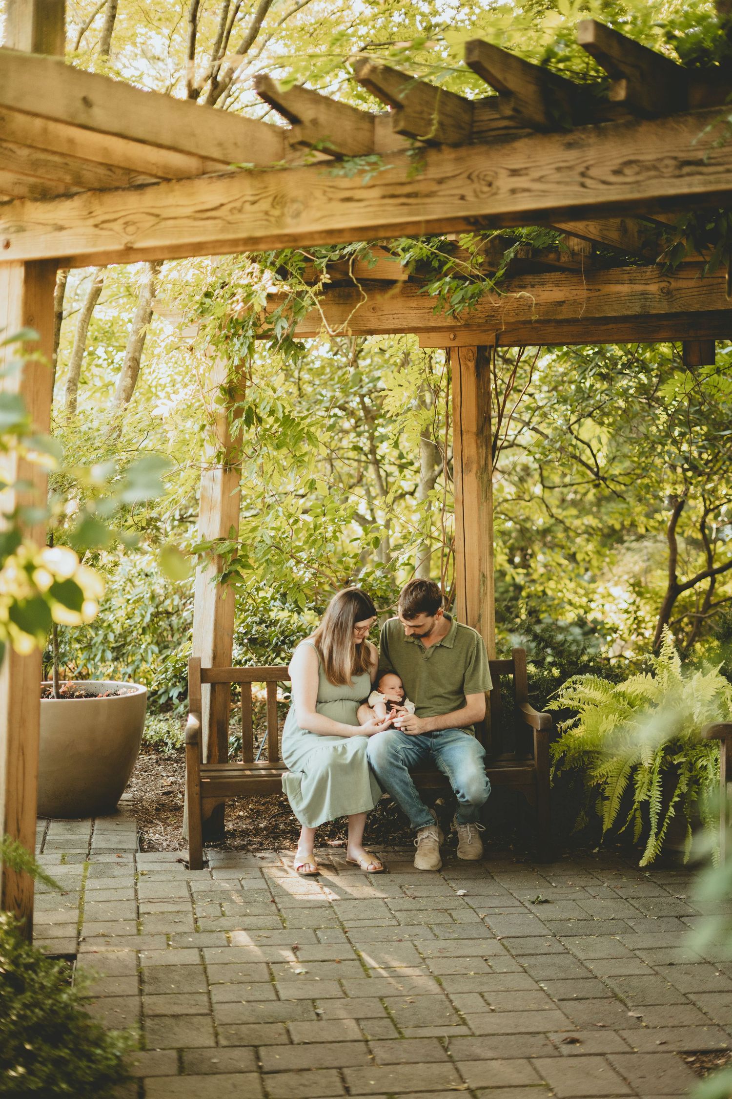 Family of three enjoys quiet moment on bench under sunlit pergola in beautiful garden setting.