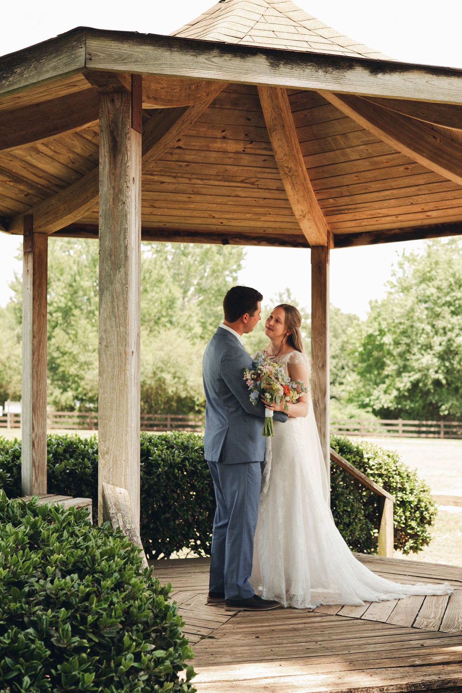 Bride and groom sharing romantic moment under wooden gazebo surrounded by lush green landscaping.