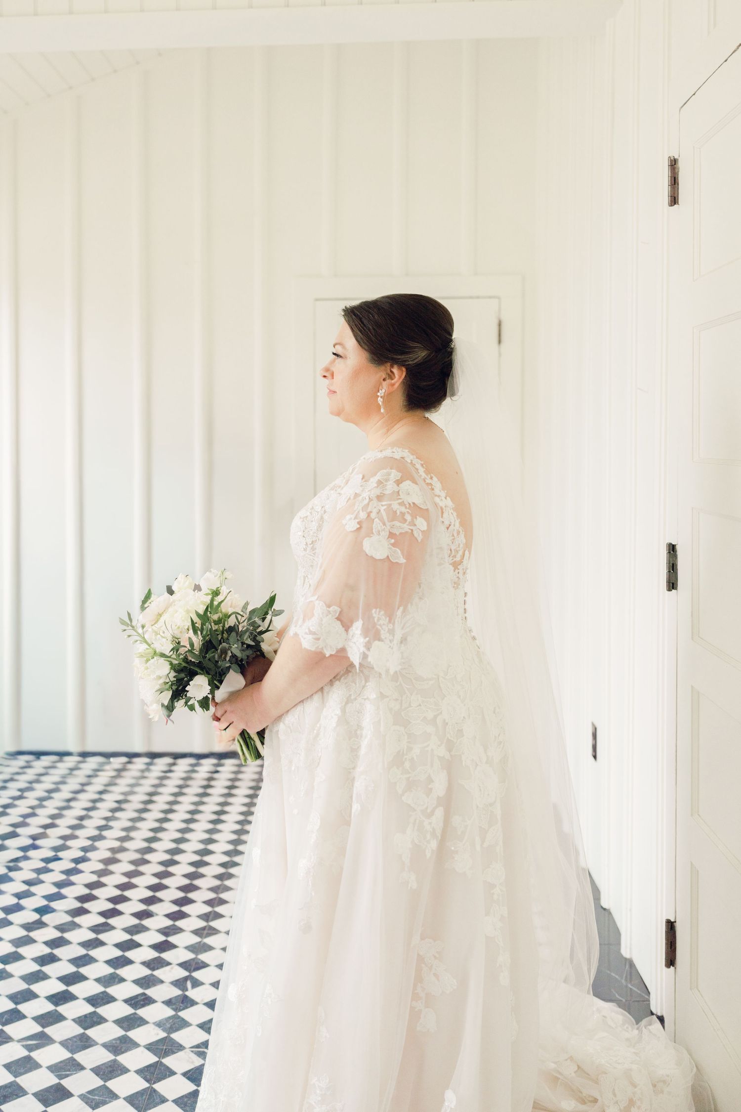 Bride in lace wedding gown holding bouquet standing in bright white room with checkered floor.
