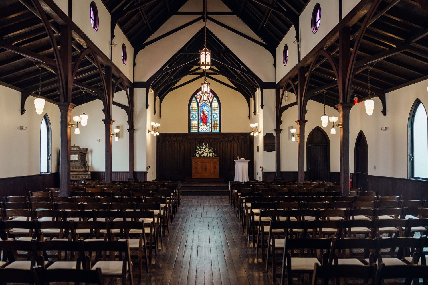 Empty historic church interior with dark wood beams, stained glass window, and rows of chairs.