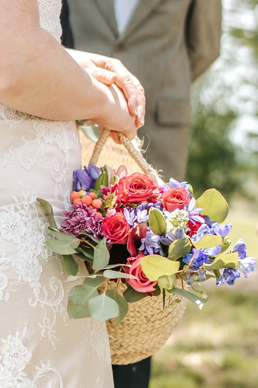 Bride holding wicker basket bouquet with colorful roses, purple flowers, and eucalyptus leaves.
