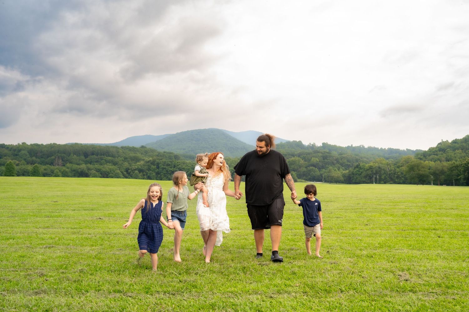 Family of five holding hands running through a green field with mountains and cloudy sky behind.