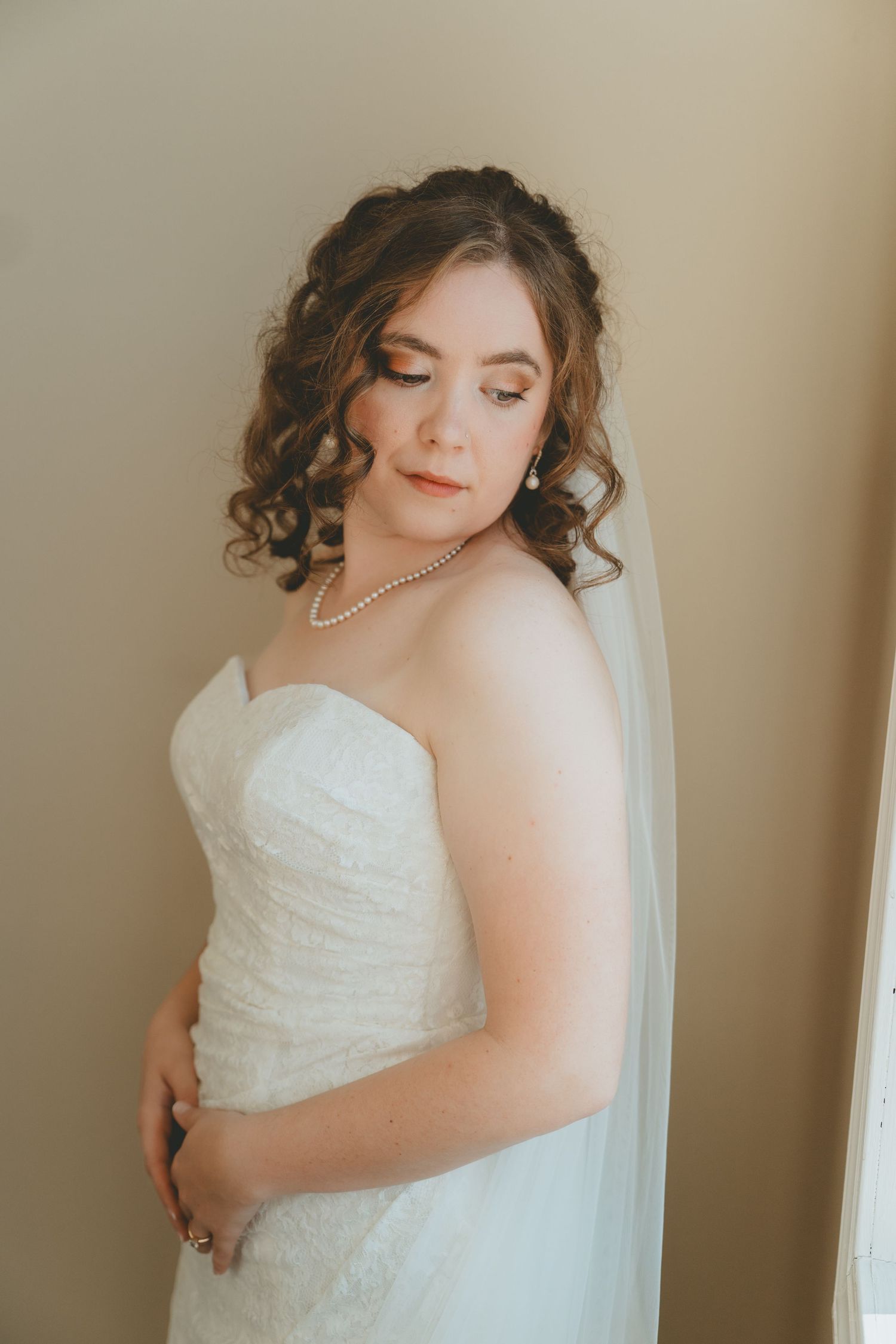 Elegant bride in strapless lace wedding gown with cathedral veil and pearl necklace poses in soft natural light.