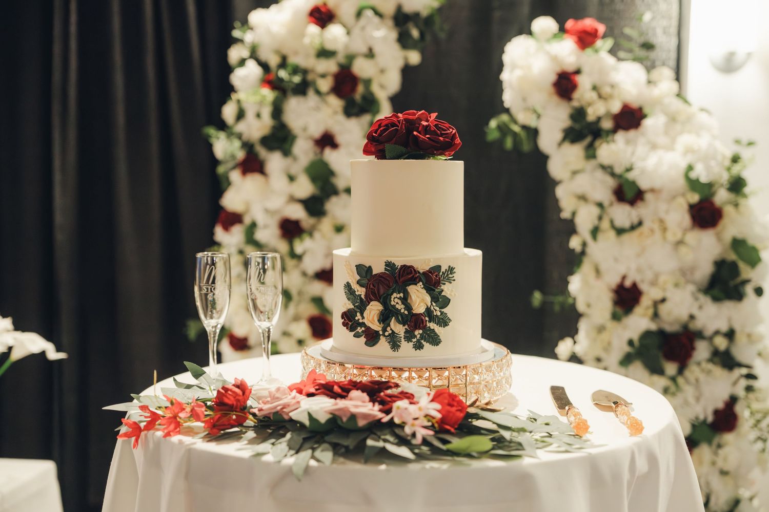 Elegant three-tier white wedding cake decorated with dark floral details, surrounded by red and white flowers on a draped table.