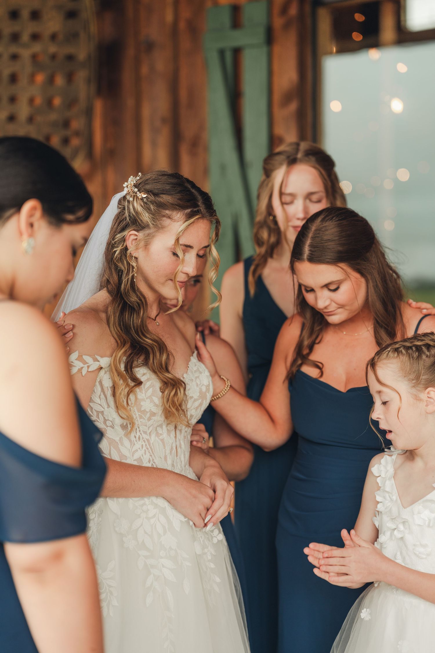 Bride in lace gown surrounded by bridesmaids in navy blue dresses and flower girl indoors.