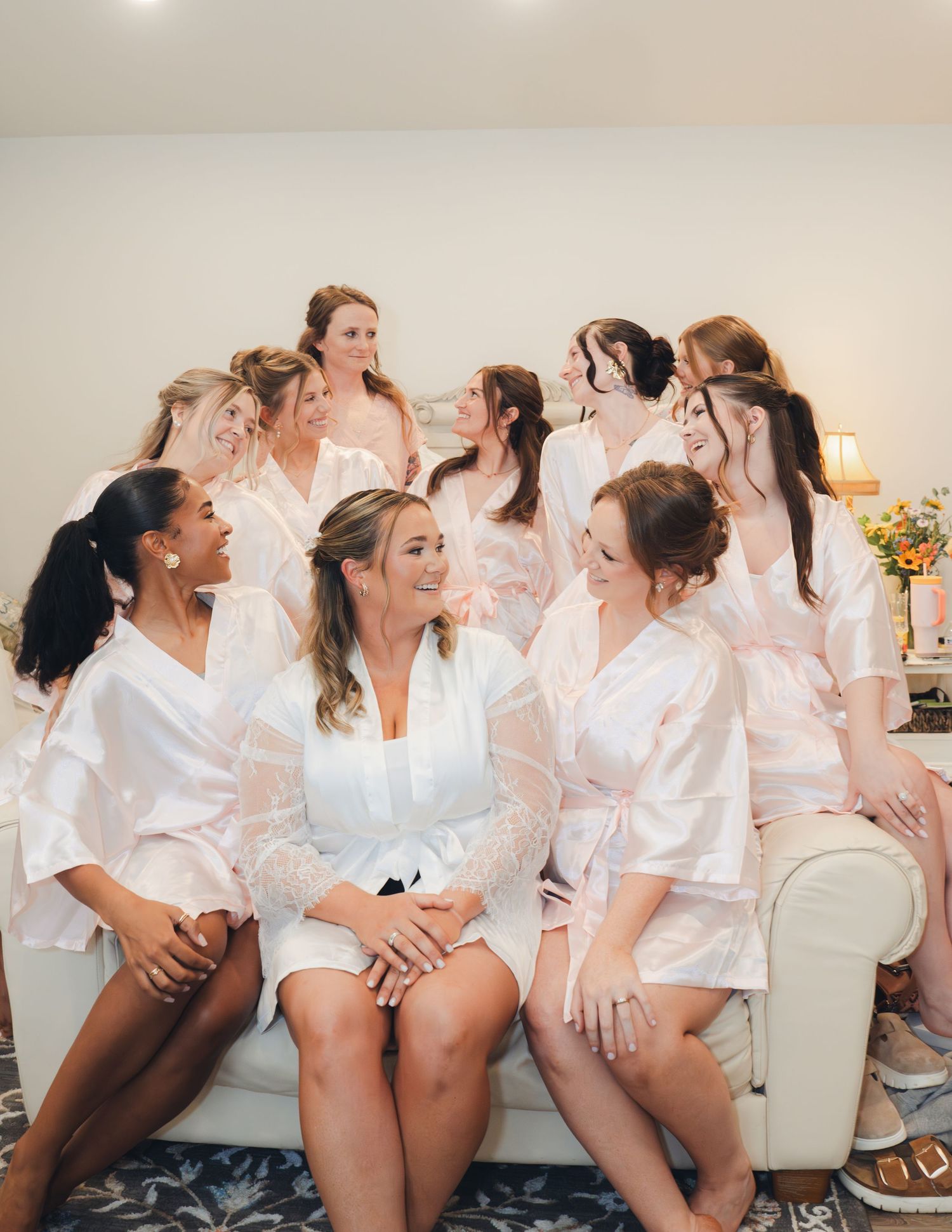 Bride in white lace robe surrounded by bridesmaids in pink robes laughing together before the wedding ceremony.