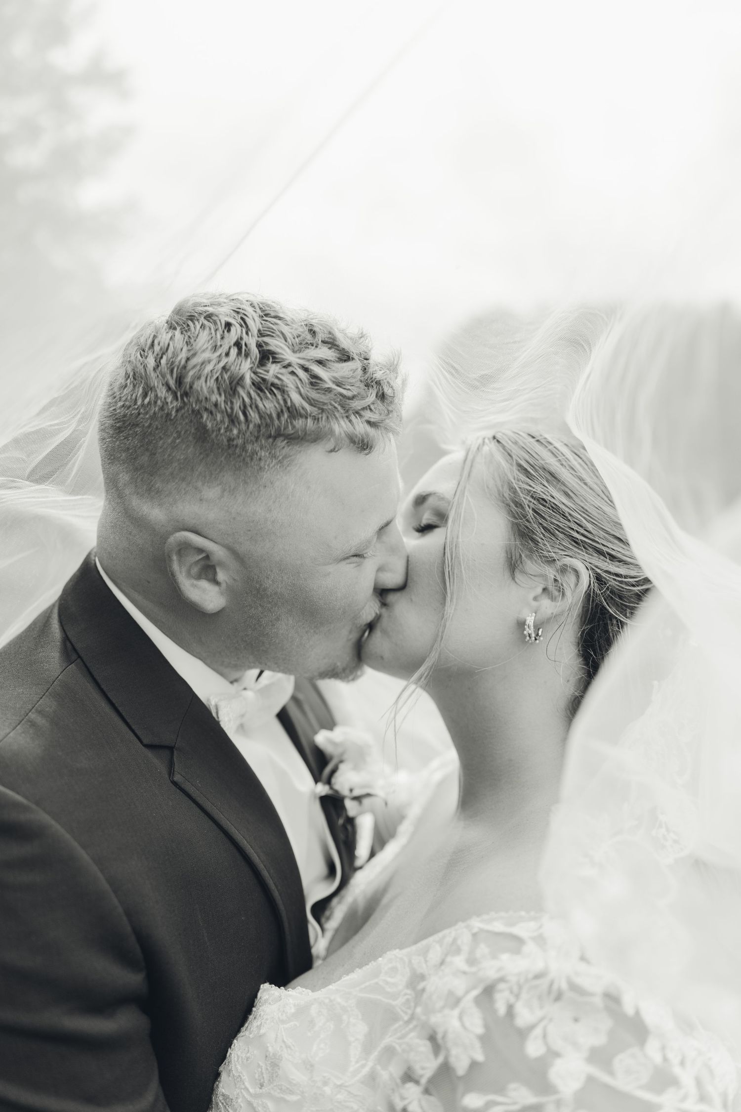 Bride and groom share a romantic kiss under a flowing veil in a dreamy black and white wedding portrait.