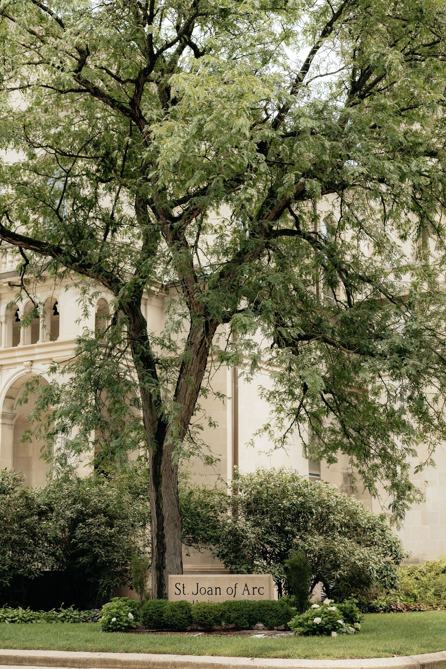 St. Joan of Arc church exterior with large green tree and stone sign in foreground.